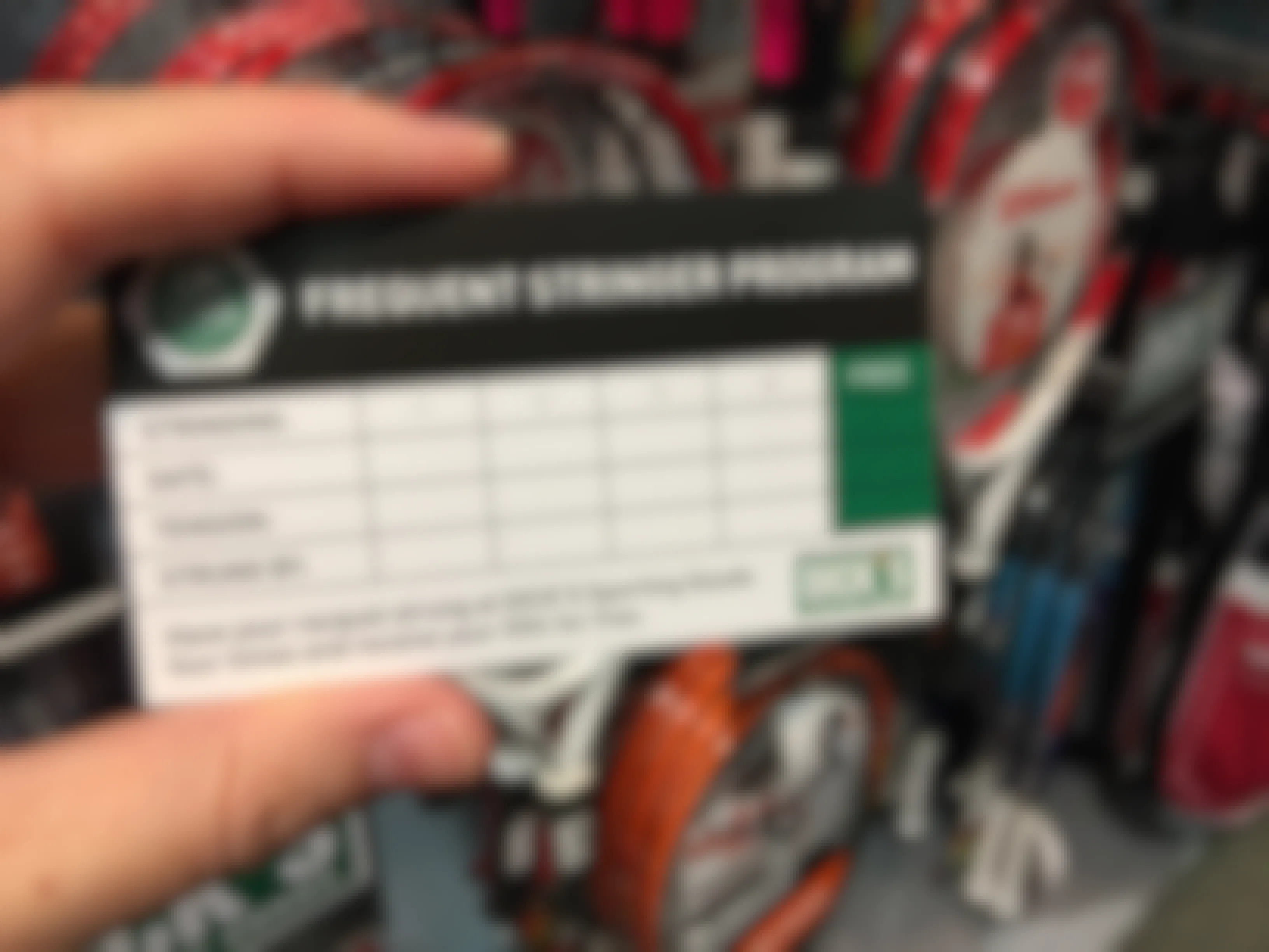 A person's hand holding up a Frequent Stringer Program card in front of a wall of rackets inside Dick's Sporting Goods.