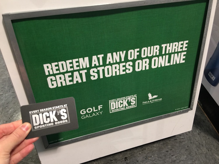 A person's hand holding a Dick's Sporting Goods gift card in front of a sign that reads "Redeem at any of our three great stores or online" and lists the stores Golf Galaxy, Dick's Sporting Goods, and Field & Stream.