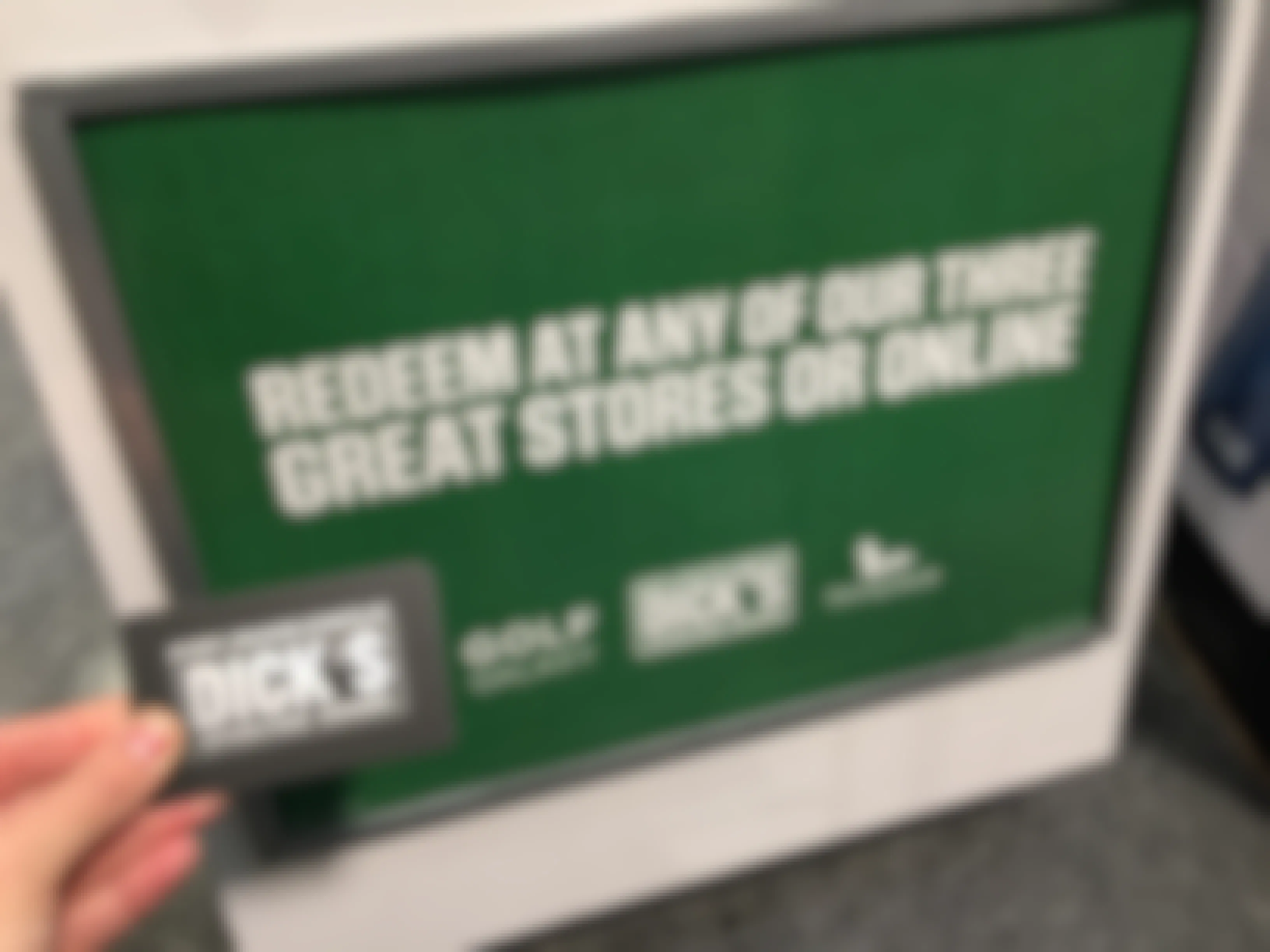 A person's hand holding a Dick's Sporting Goods gift card in front of a sign that reads "Redeem at any of our three great stores or online" and lists the stores Golf Galaxy, Dick's Sporting Goods, and Field & Stream.