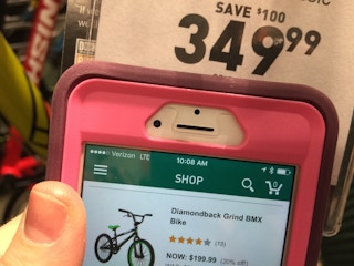 A close up of a price tag on a bike with a lower price pulled up on the Dick's app for the same item.