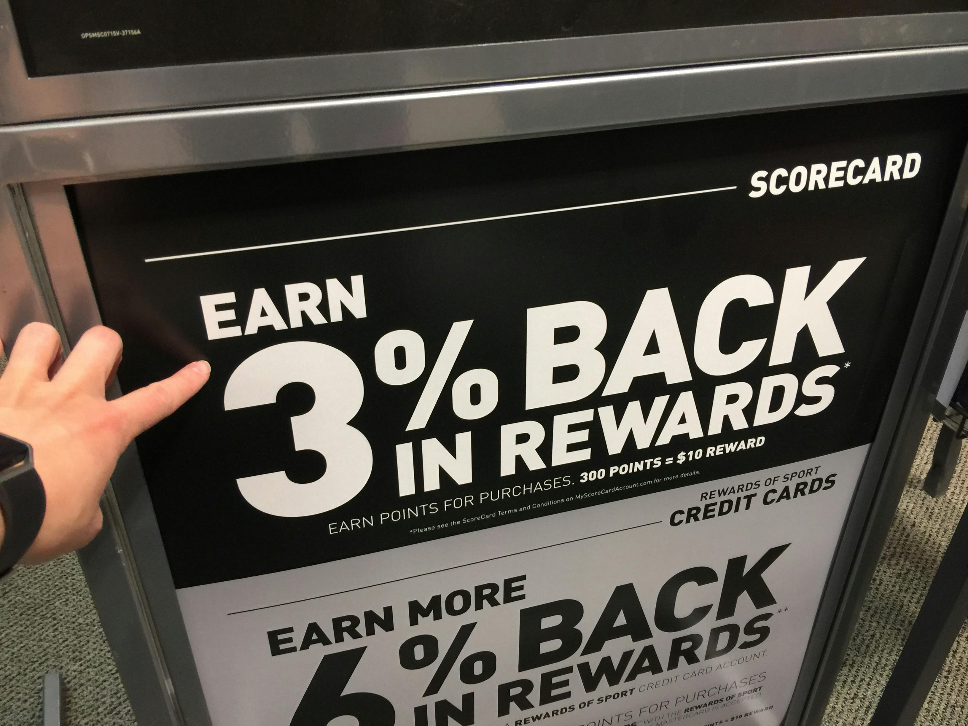 A person's hand pointing to a sign inside Dick's Sporting Goods advertising the 3% back in rewards you can get with ScoreCard program.