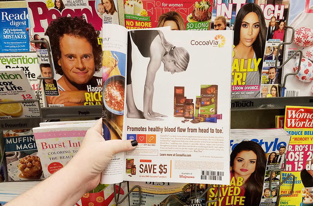 A person holding a magazine open to a coupon in front of a magazine stand with other magazines.