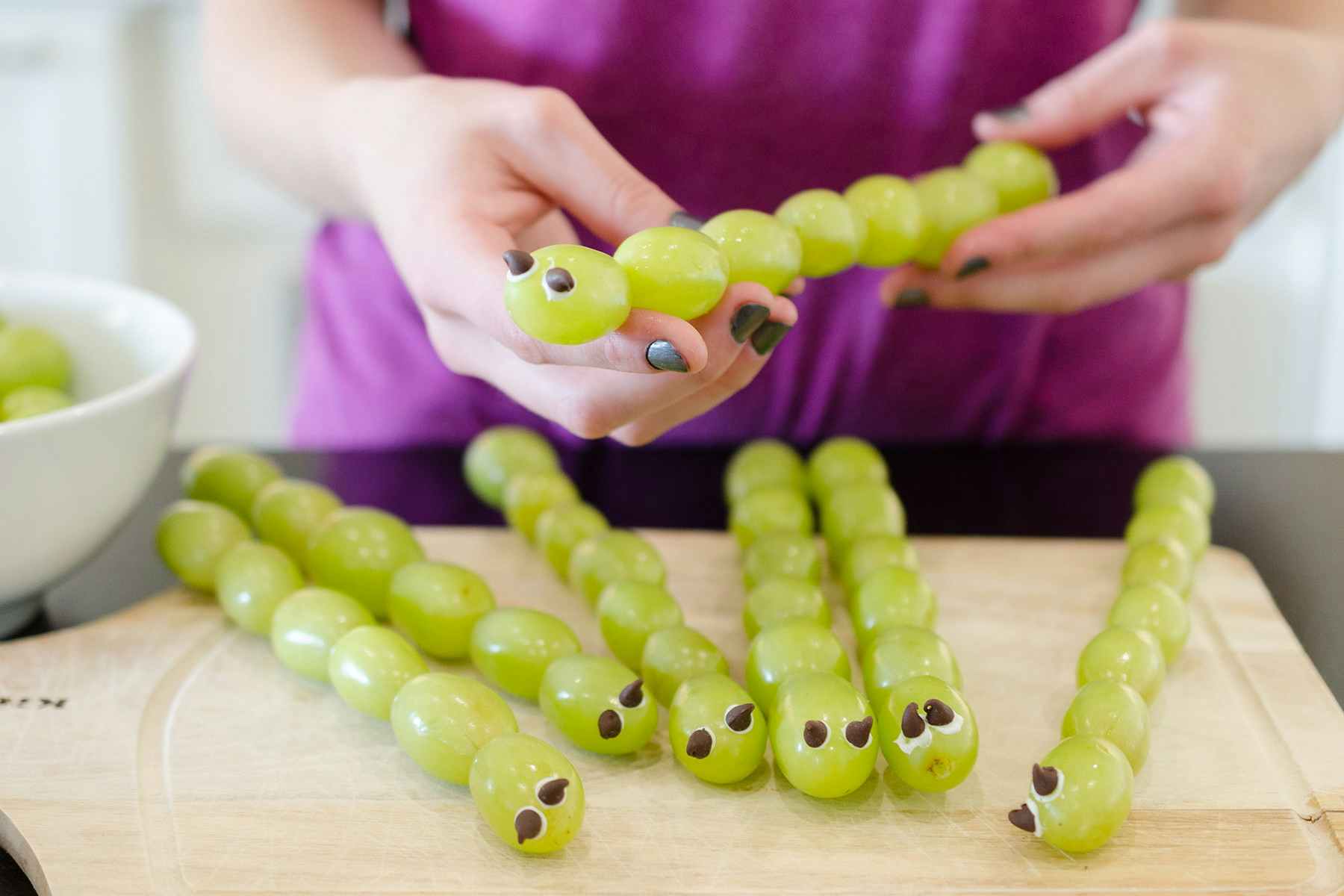 A woman holding a snack made of grapes and chocolate chips to look like a caterpillar.