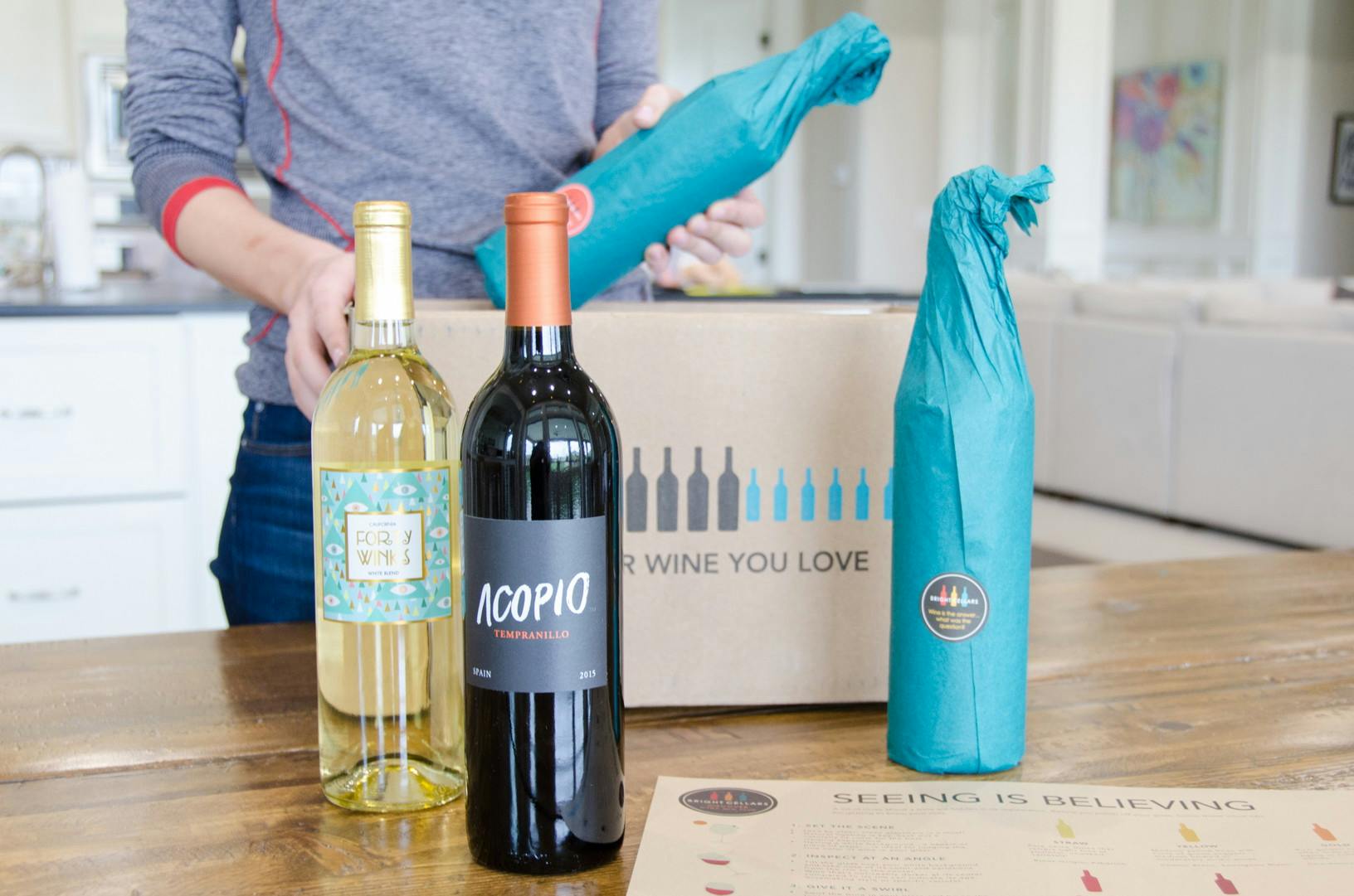 Woman holding a bottle of wine above a box with three wines bottles standing in front of the box.