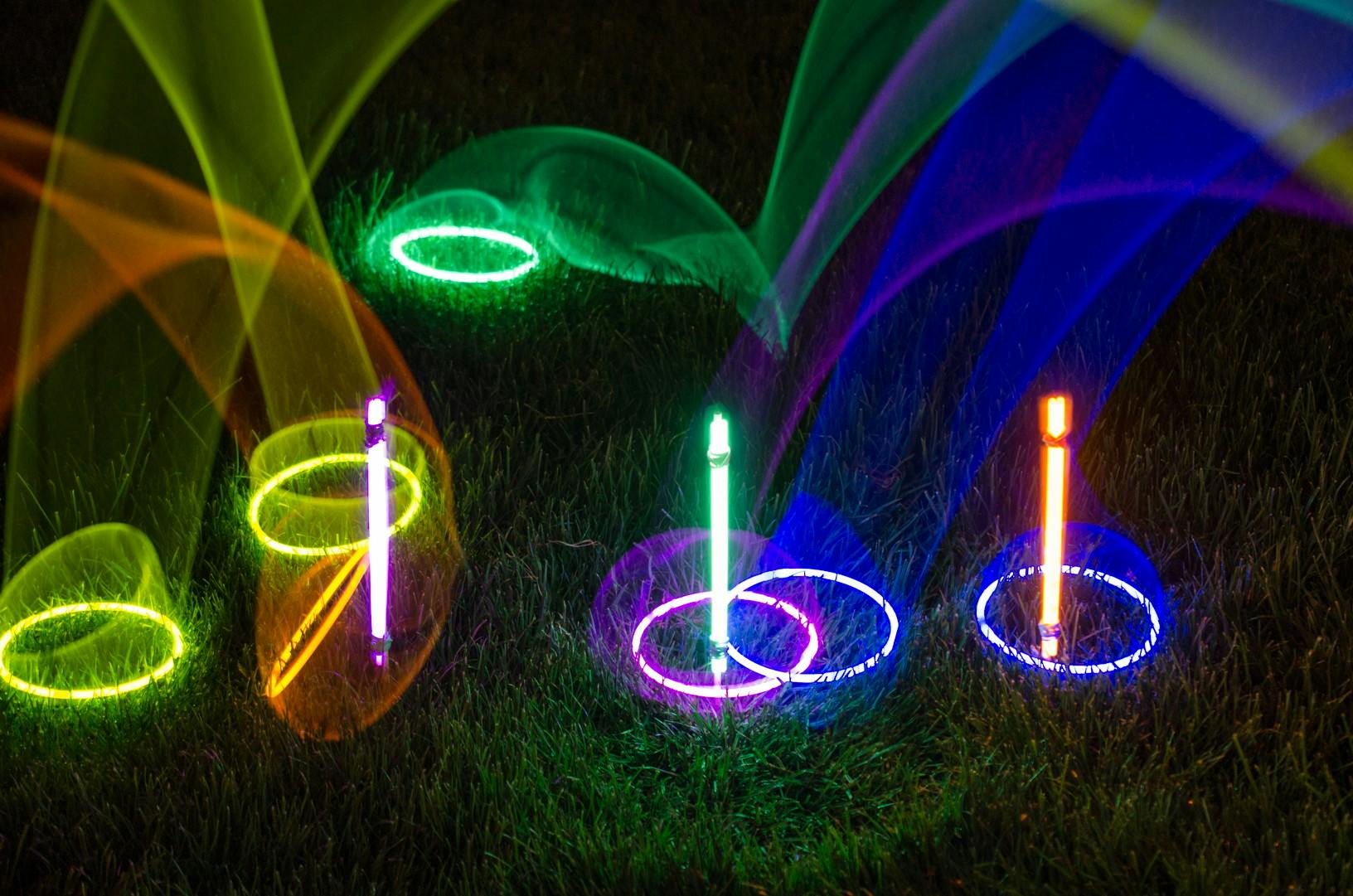 Glow stick necklaces being used in a ring toss game at night, with glow sticks sticking out of the ground as targets.