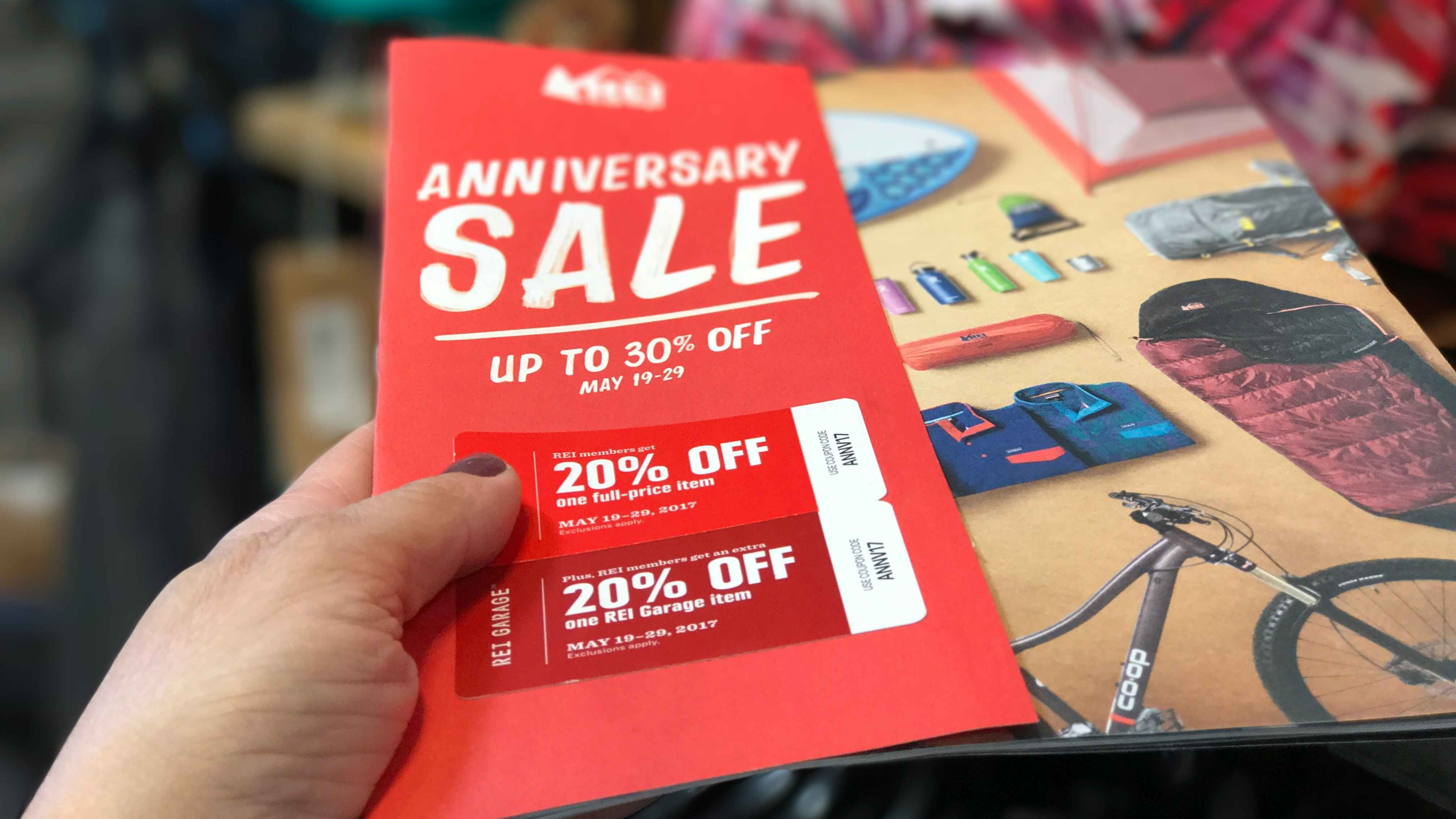 A person's hand holding up some REI Anniversary Sale coupons above a REI ad book.