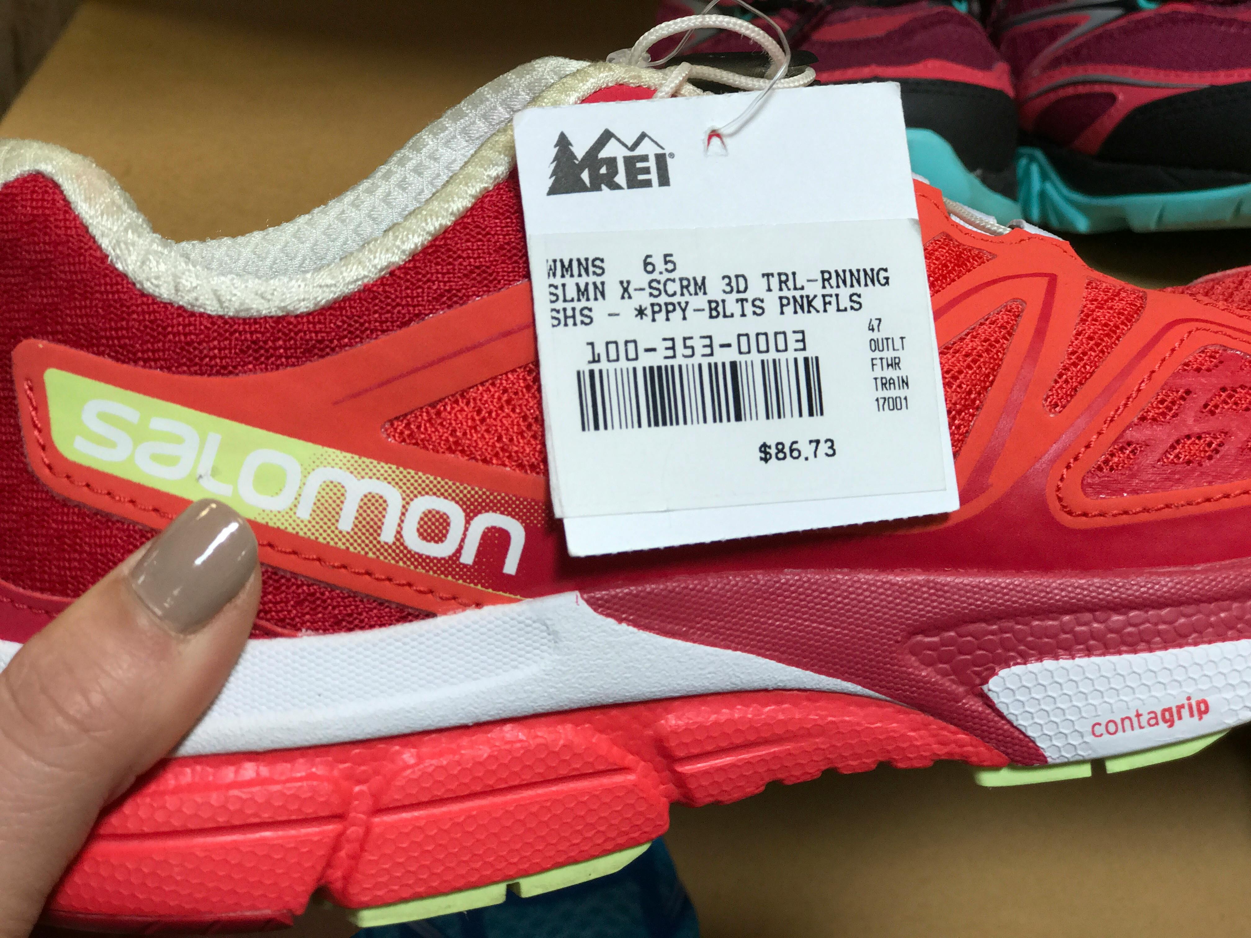 A close up of a person's hand holding a Salomon sneaker and showing the tag at REI.