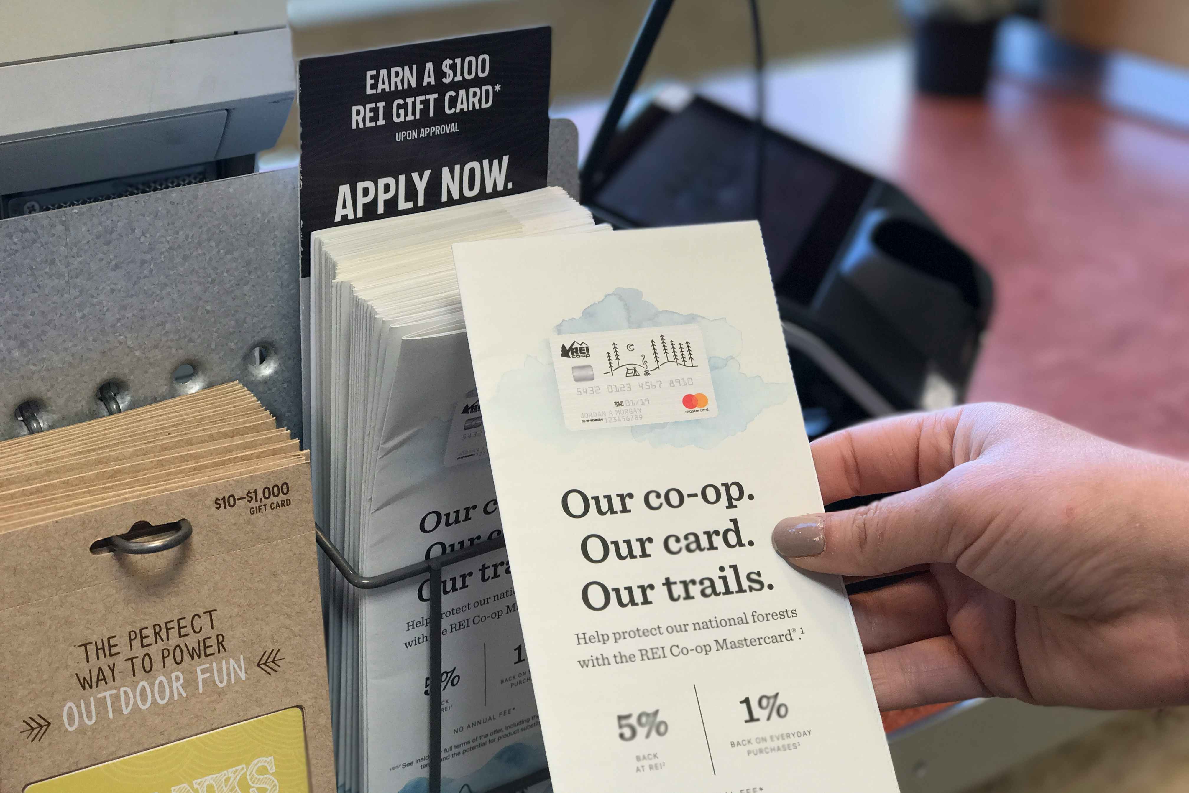 A person's hand taking a brochure for the REI credit card from a display at the REI checkout counter.