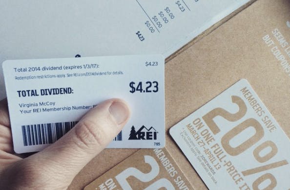 A person's hand holding a dividend card from REI with $4.23 on it, next to some members coupons from REI.