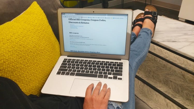 A person sitting on a couch with their laptop open on their lap, browsing the REI promotions page on their website.