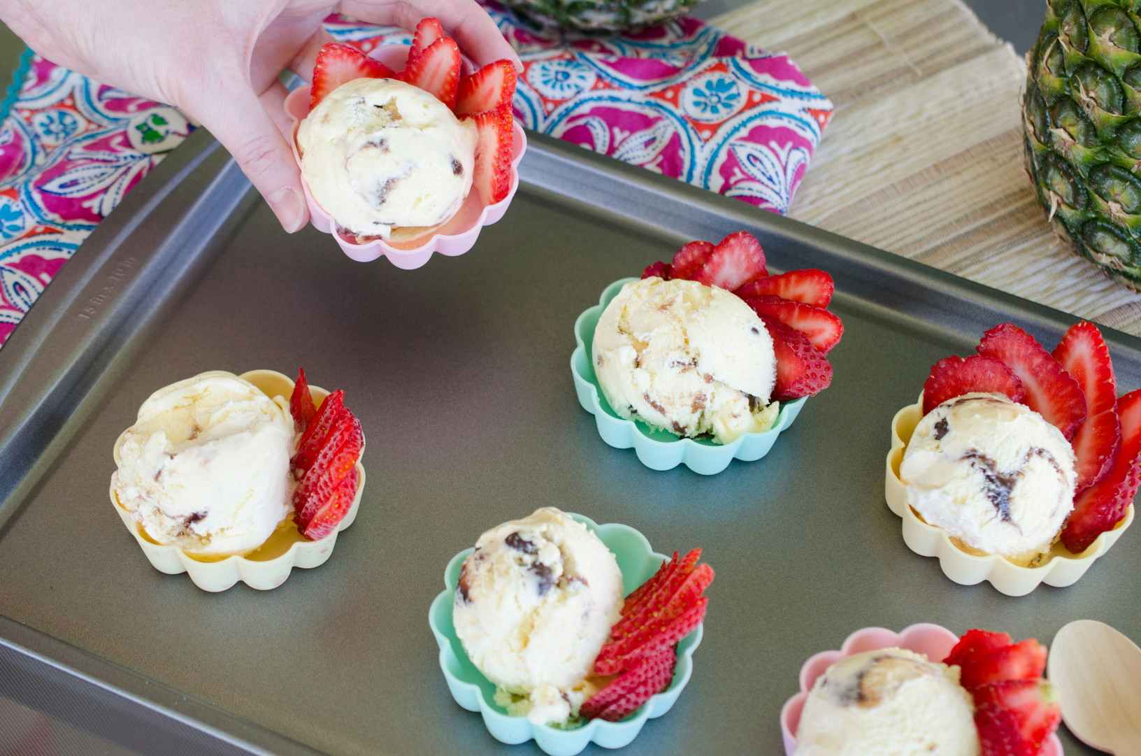 A person's hand picking up a cup of ice cream from a tray of little colorful cups with pre-scooped ice cream and strawberries.