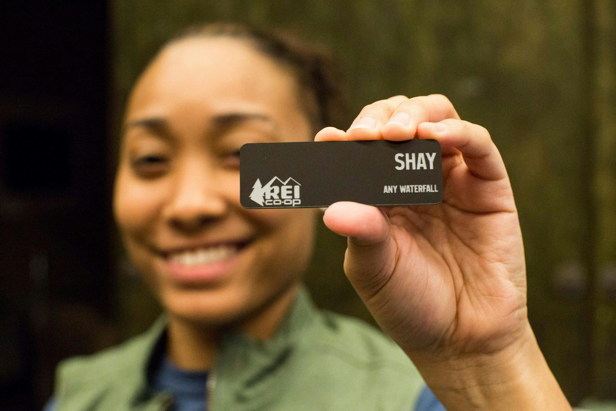 A woman holding up an REI employee name tag that reads "Shay