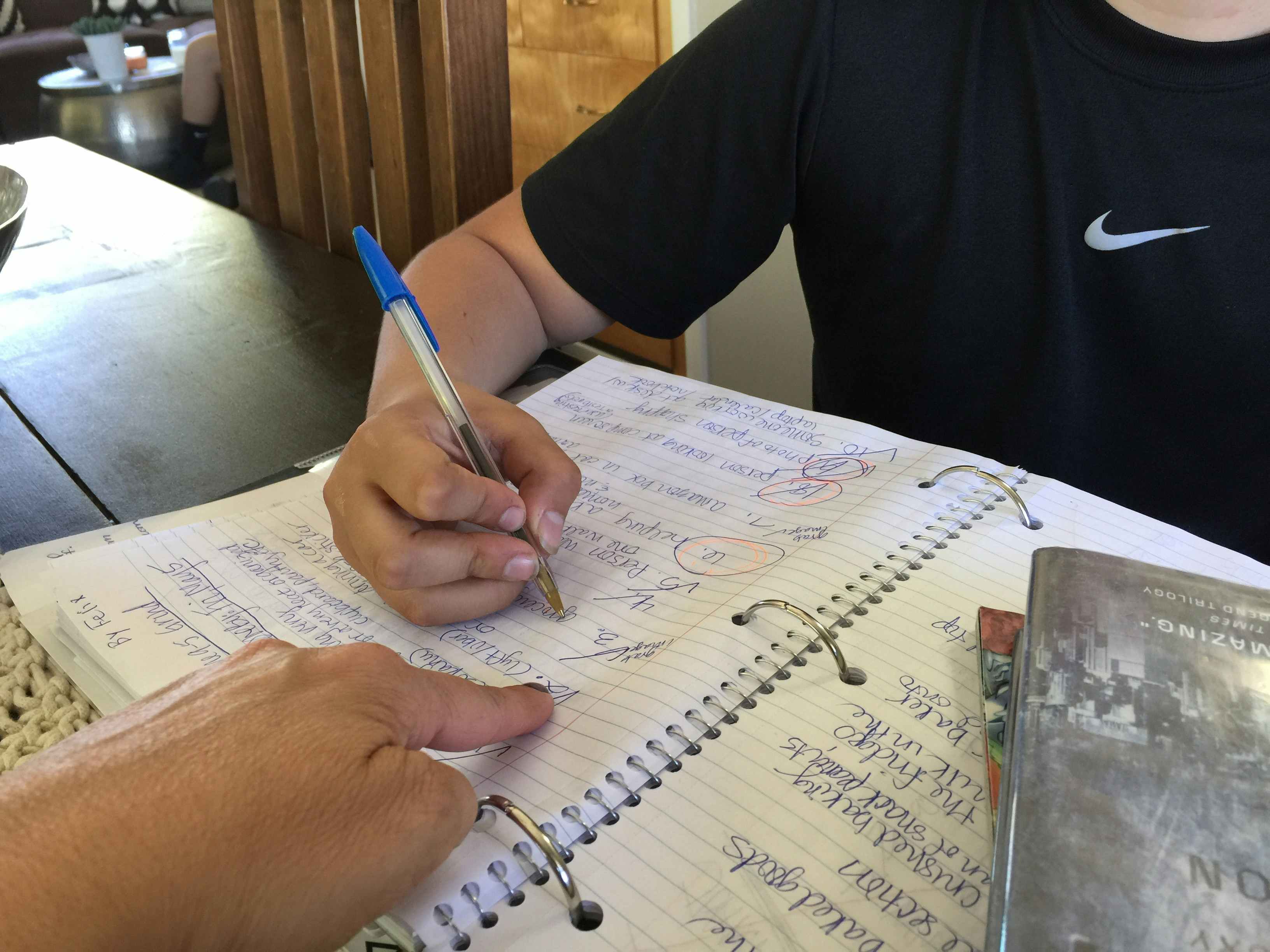 A tutor pointing to a line of a student's homework as he is writing in a notebook.