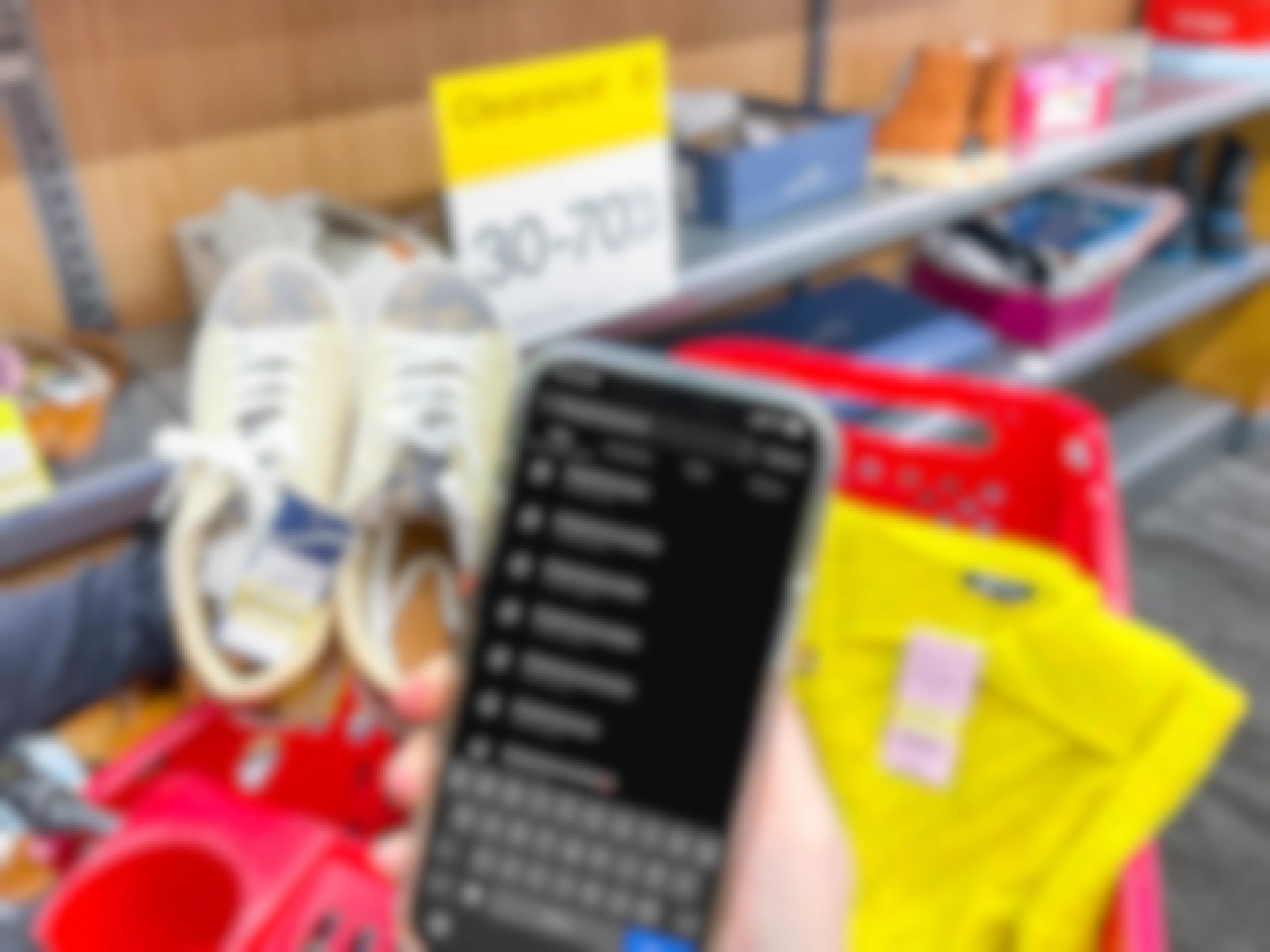 A person's hand holding up a cellphone displaying the search results for #targetclearance on Instagram in front of the clearance shoe section at Target and a sign advertising clearance as 30-70% off.