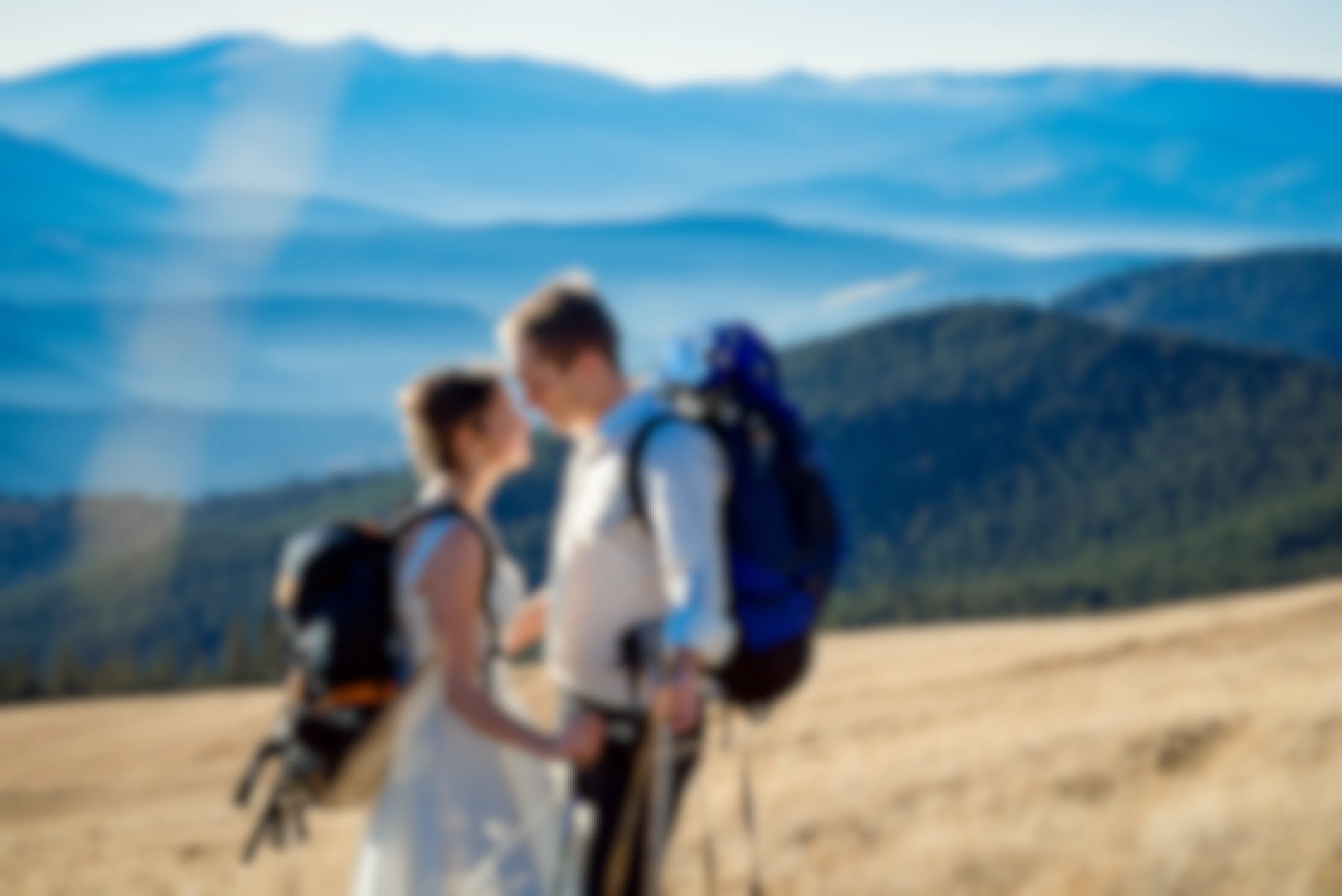 A man and woman standing on a mountain side with mountains in the distance behind them. The woman is dressed in a wedding gown and the man is wearing slacks and a dress shirt. Both are wearing hiking backpacks and carrying hiking polls.