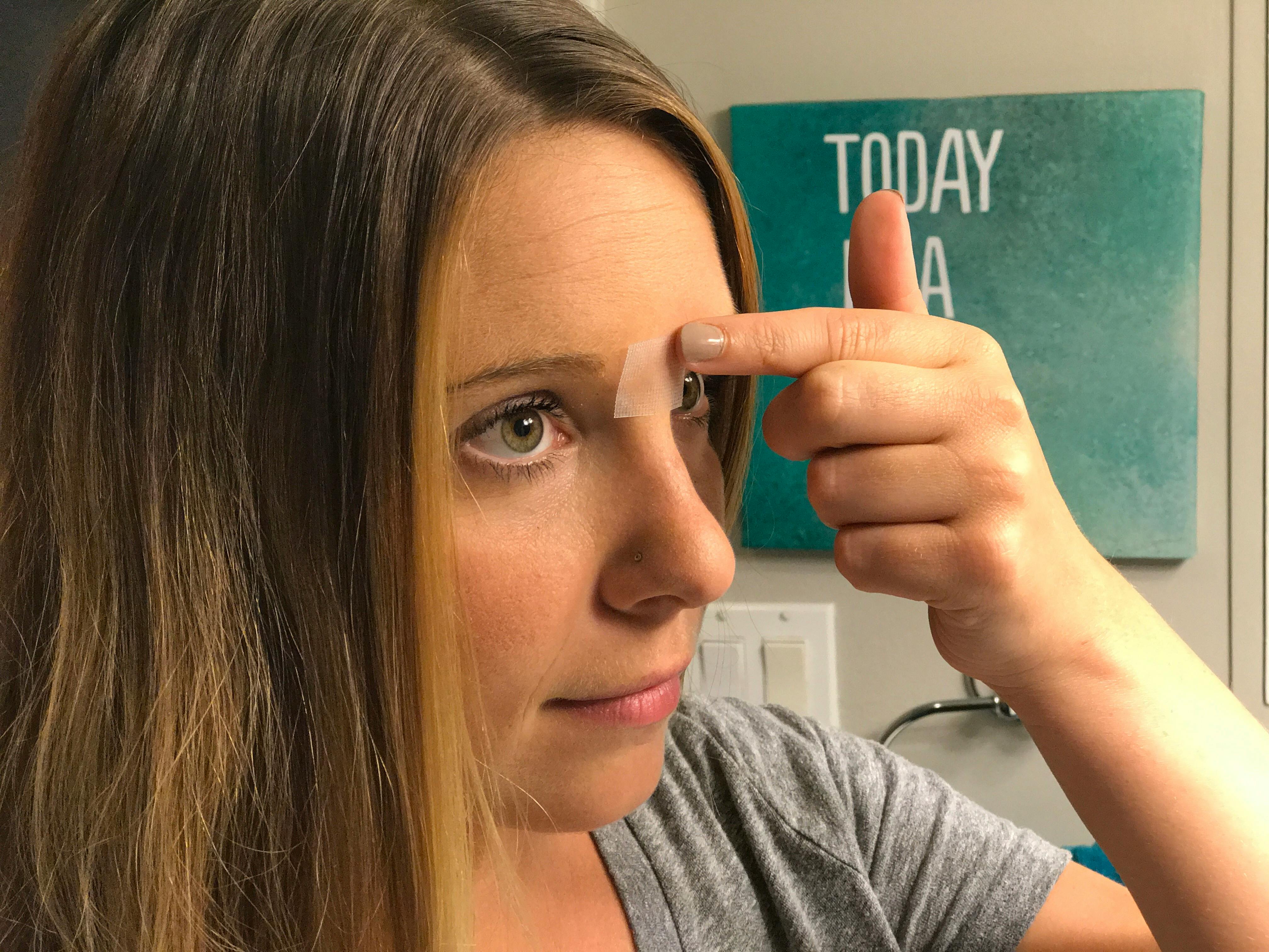 A woman putting tape between her eyebrows just above the bridge of her nose