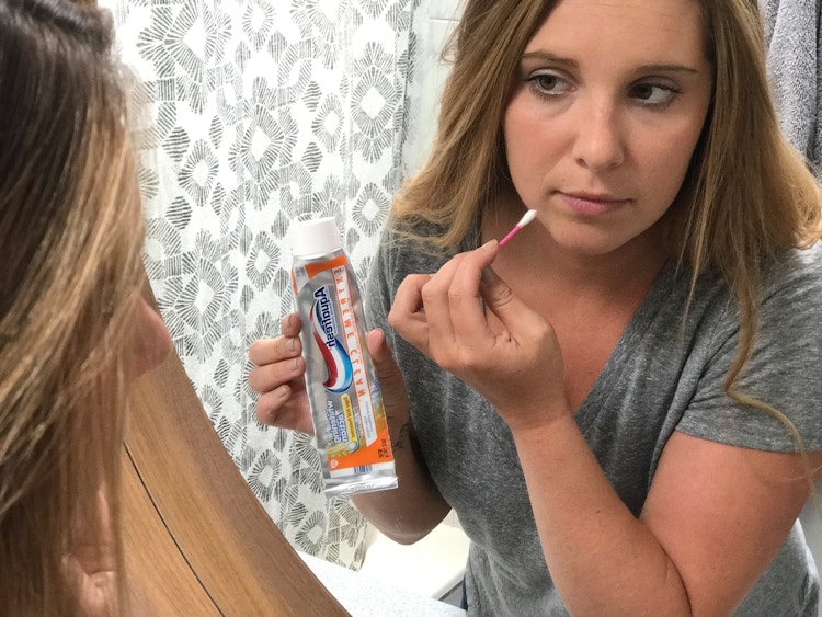 A woman using a Q-tip to apply toothpaste to a blemish on her face