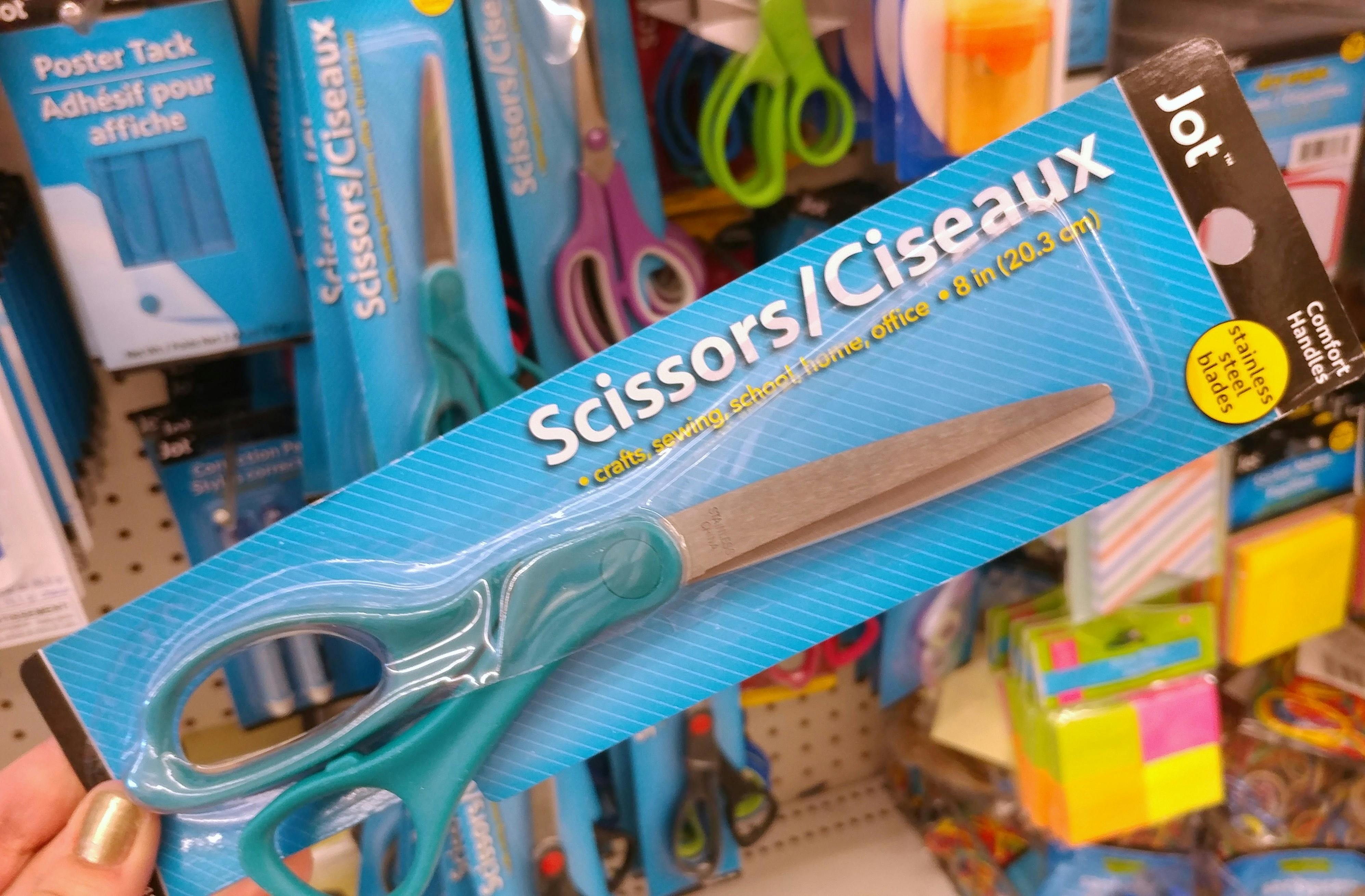 A person's hand holding up a pair of scissors in front of the display of scissors for sale at Dollar Tree.