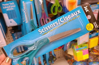 A person's hand holding up a pair of scissors in front of the display of scissors for sale at Dollar Tree.