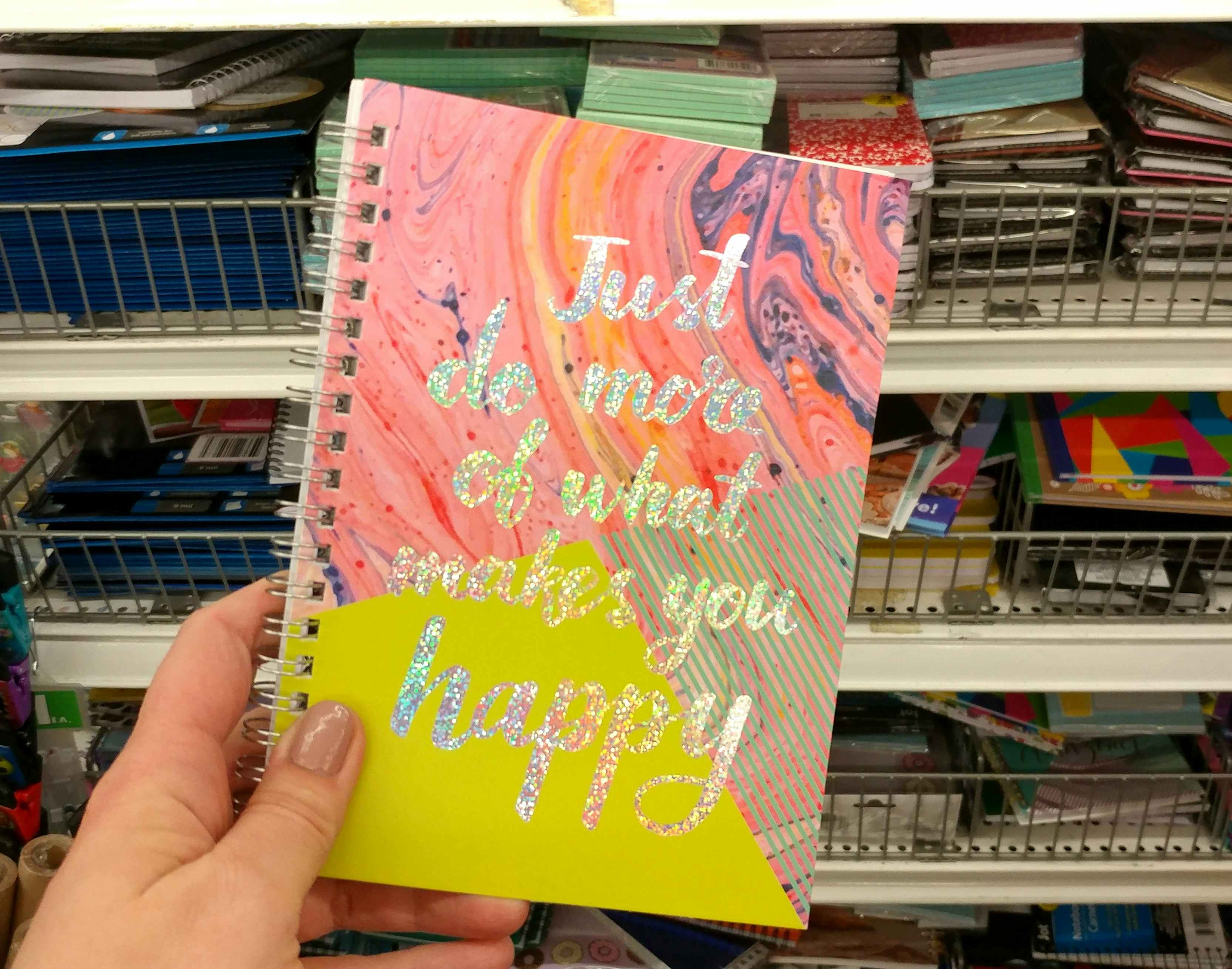 A person's hand holding a colorful spiral notebook in front of a shelf of more notebooks at Dollar Tree.