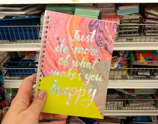 A person's hand holding a colorful spiral notebook in front of a shelf of more notebooks at Dollar Tree.