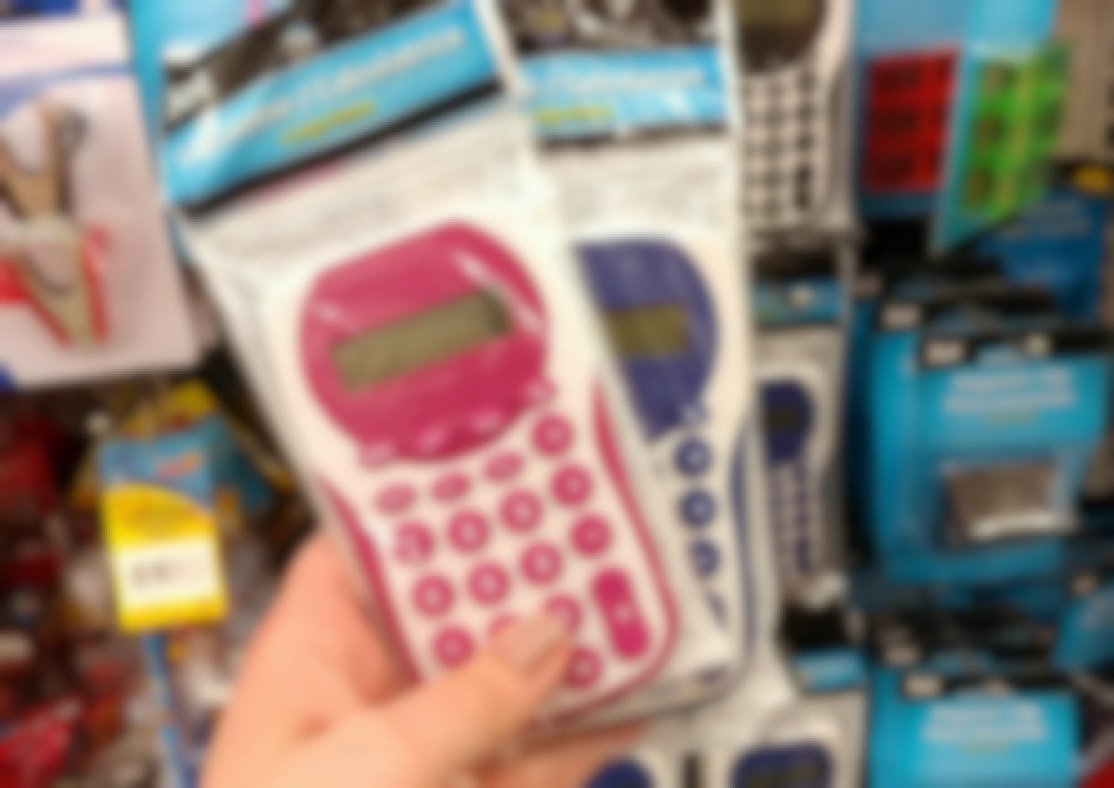 A person's hand holding up two calculators in front of a display of calculators for sale at Dollar Tree.