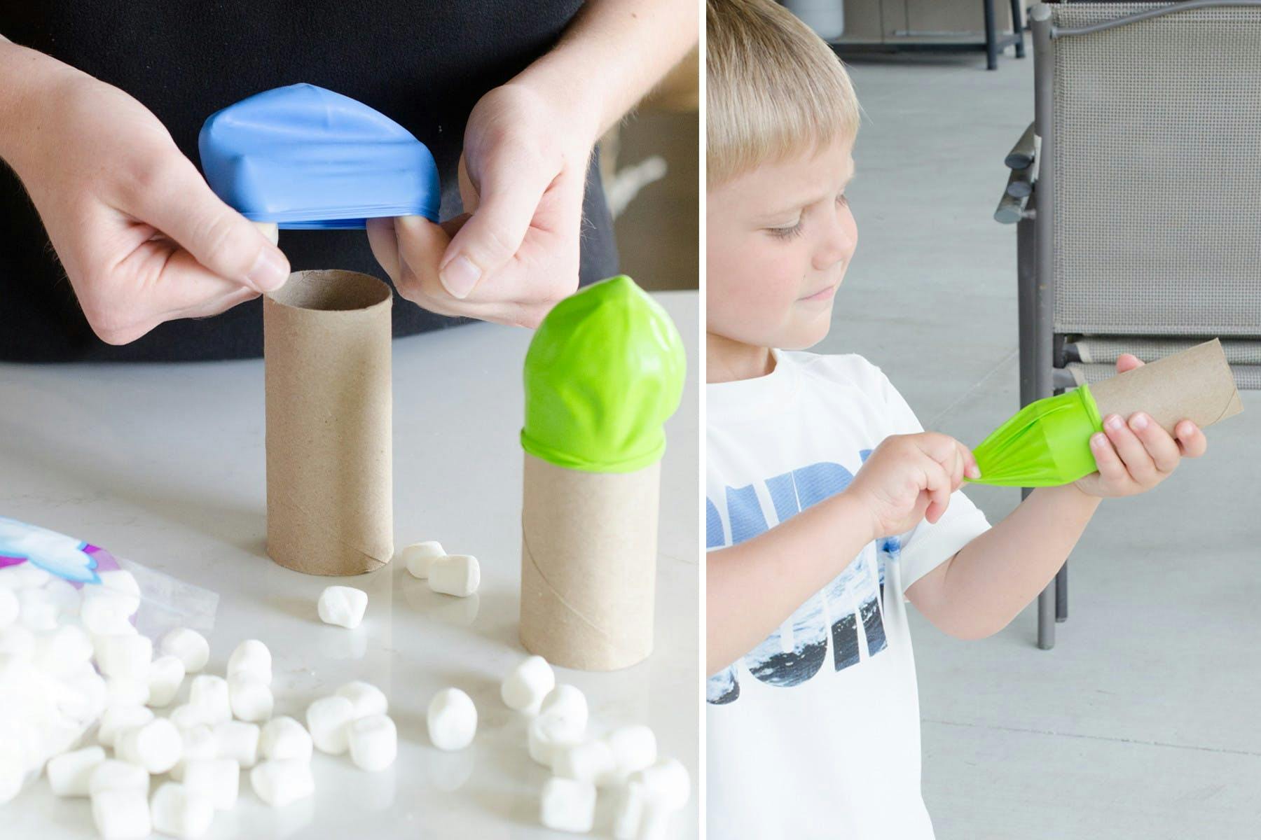 Someone putting a balloon on the end of a toilet paper roll next to some marshmallows on a table, and a child using the contraption to shoot marshmallows
