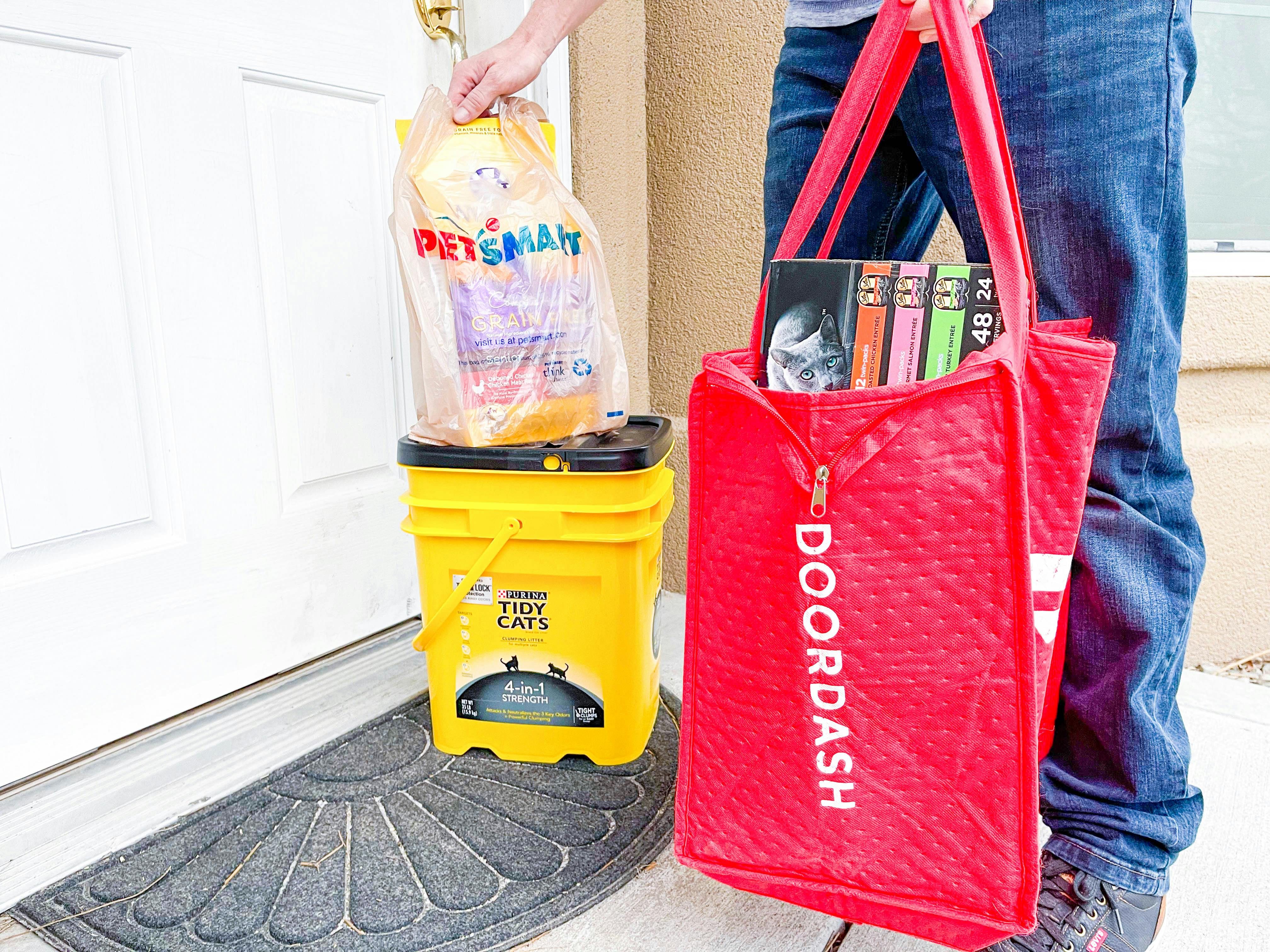 A Doordash delivery person standing on a front porch, setting a PetSmart bag onto a box of Tidy Cat kitty litter and holding a large DoorDash delivery bag with more pet products.