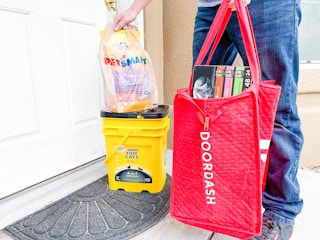 A Doordash delivery person standing on a front porch, setting a PetSmart bag onto a box of Tidy Cat kitty litter and holding a large DoorDash delivery bag with more pet products.