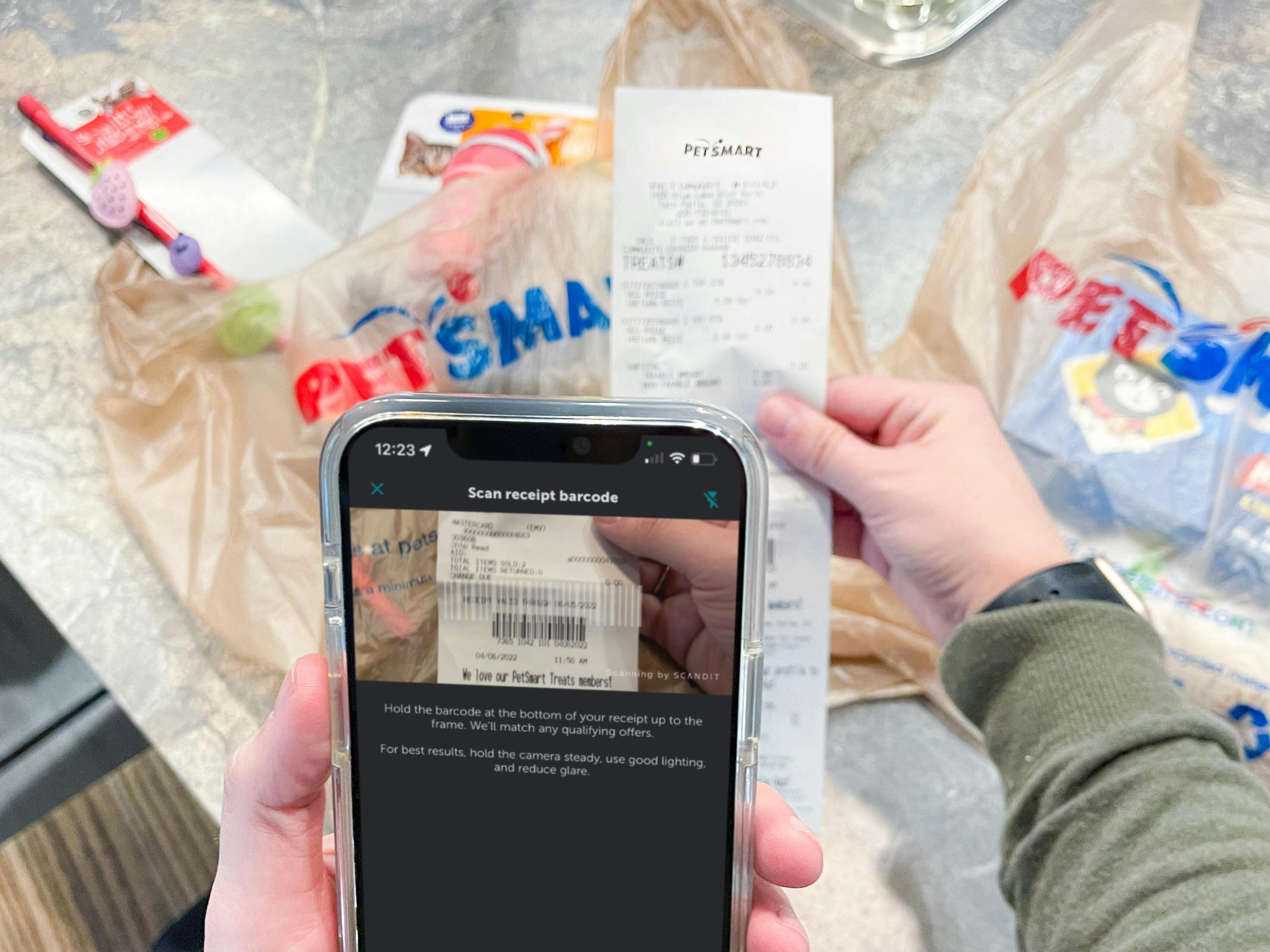A person's hands holding up a PetSmart receipt in front of two PetSmart bags and using the Ibotta receipt scanning feature on their cell phone to scan the receipt.