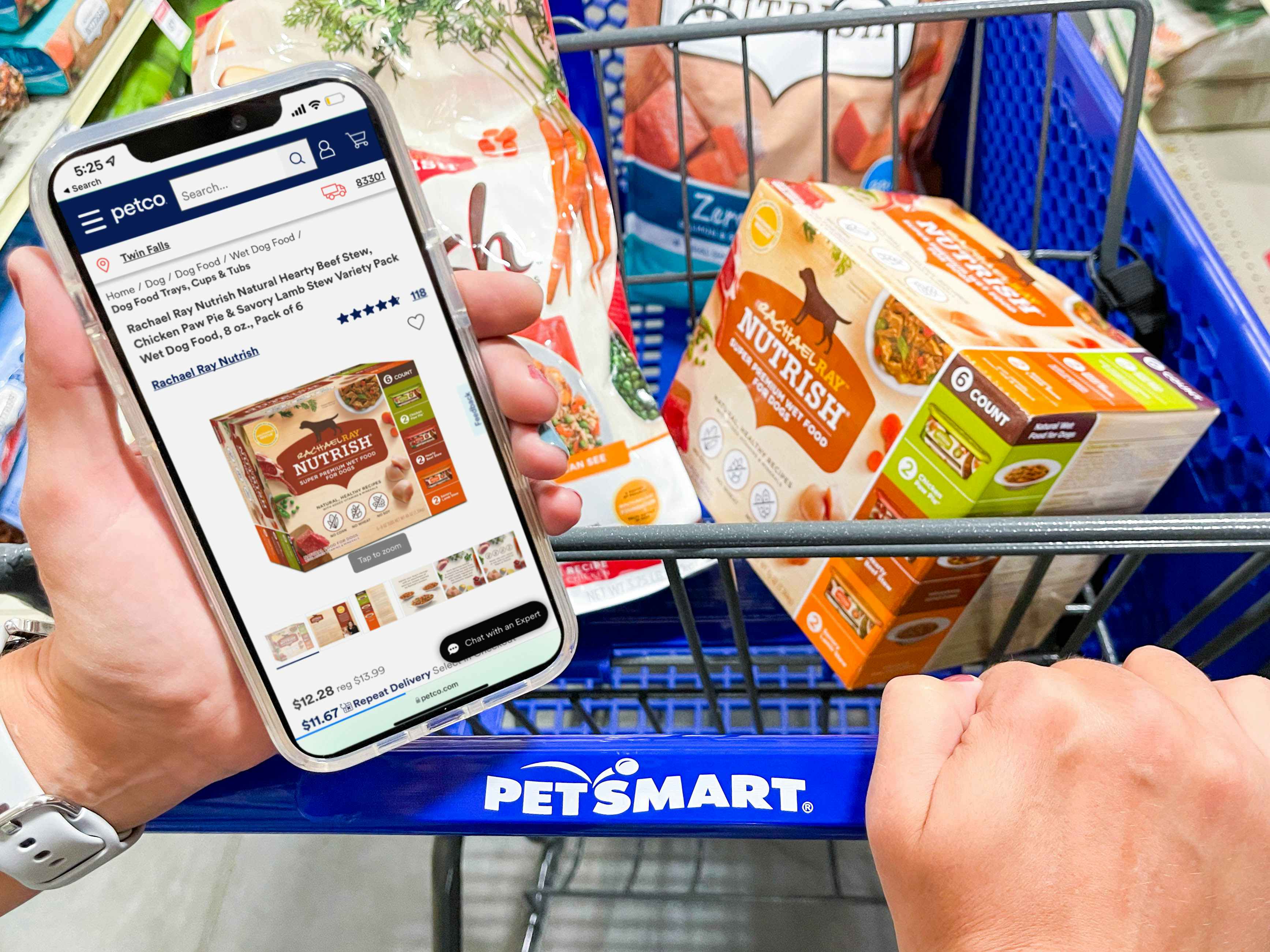 A person's hands, one holding a cellphone with the Petco website open to a product, and the other hand pushing a PetSmart shopping cart with that product in the basket.