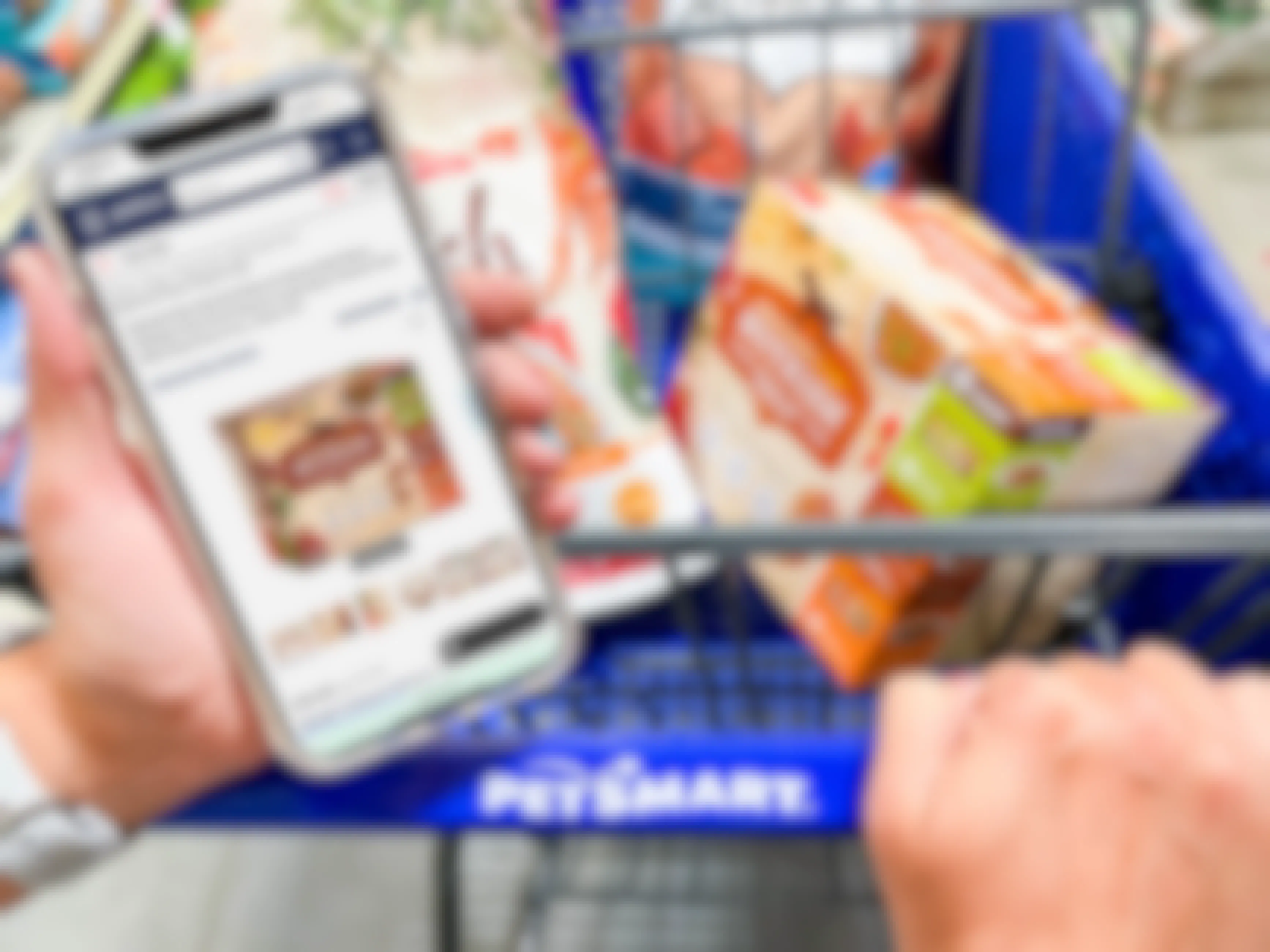 A person's hands, one holding a cellphone with the Petco website open to a product, and the other hand pushing a PetSmart shopping cart with that product in the basket.