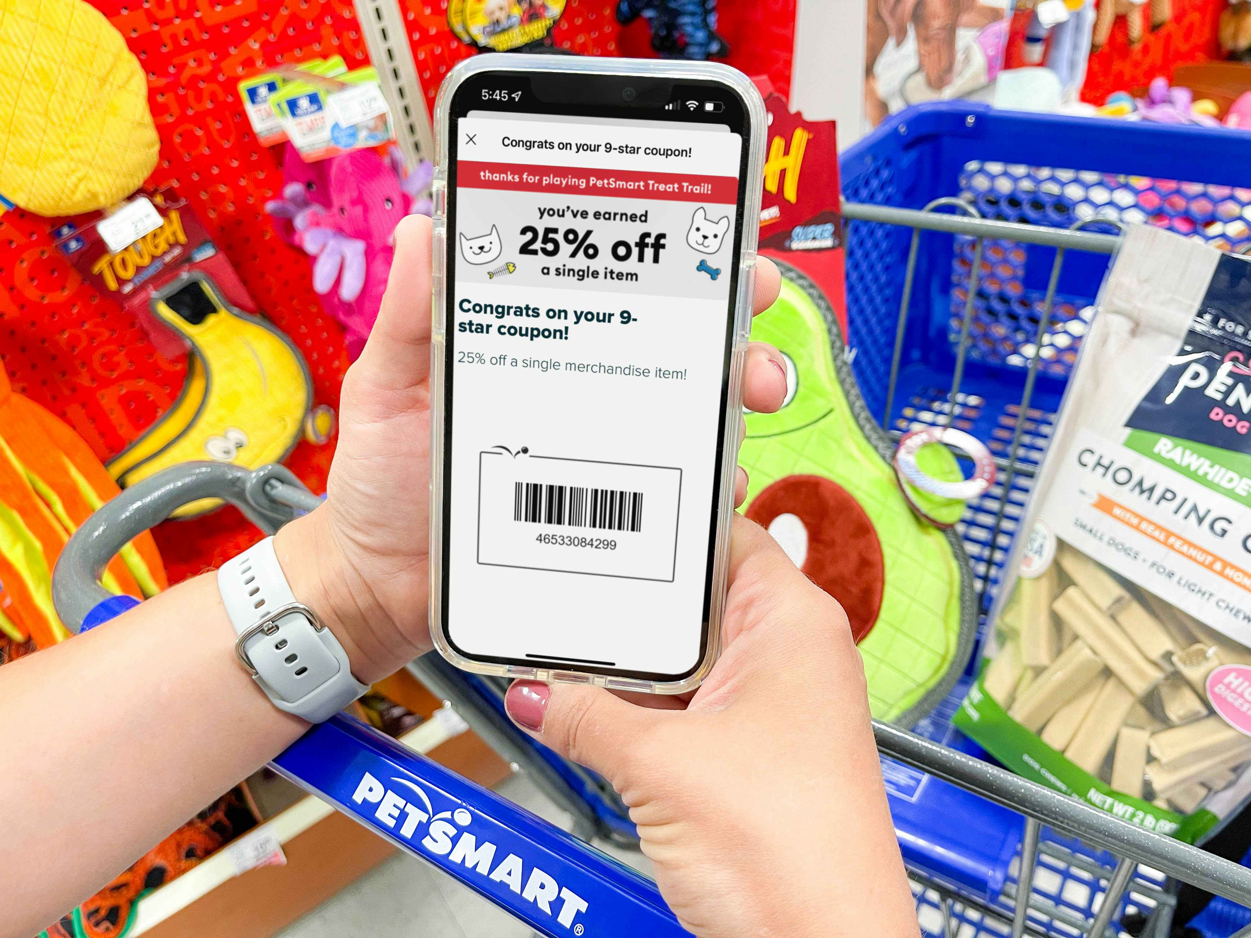 A person's hands holding a cellphone displaying a Treat Trail game coupon on the Petsmart app in front of a PetSmart shopping cart with a dog toy and treats in the basket.