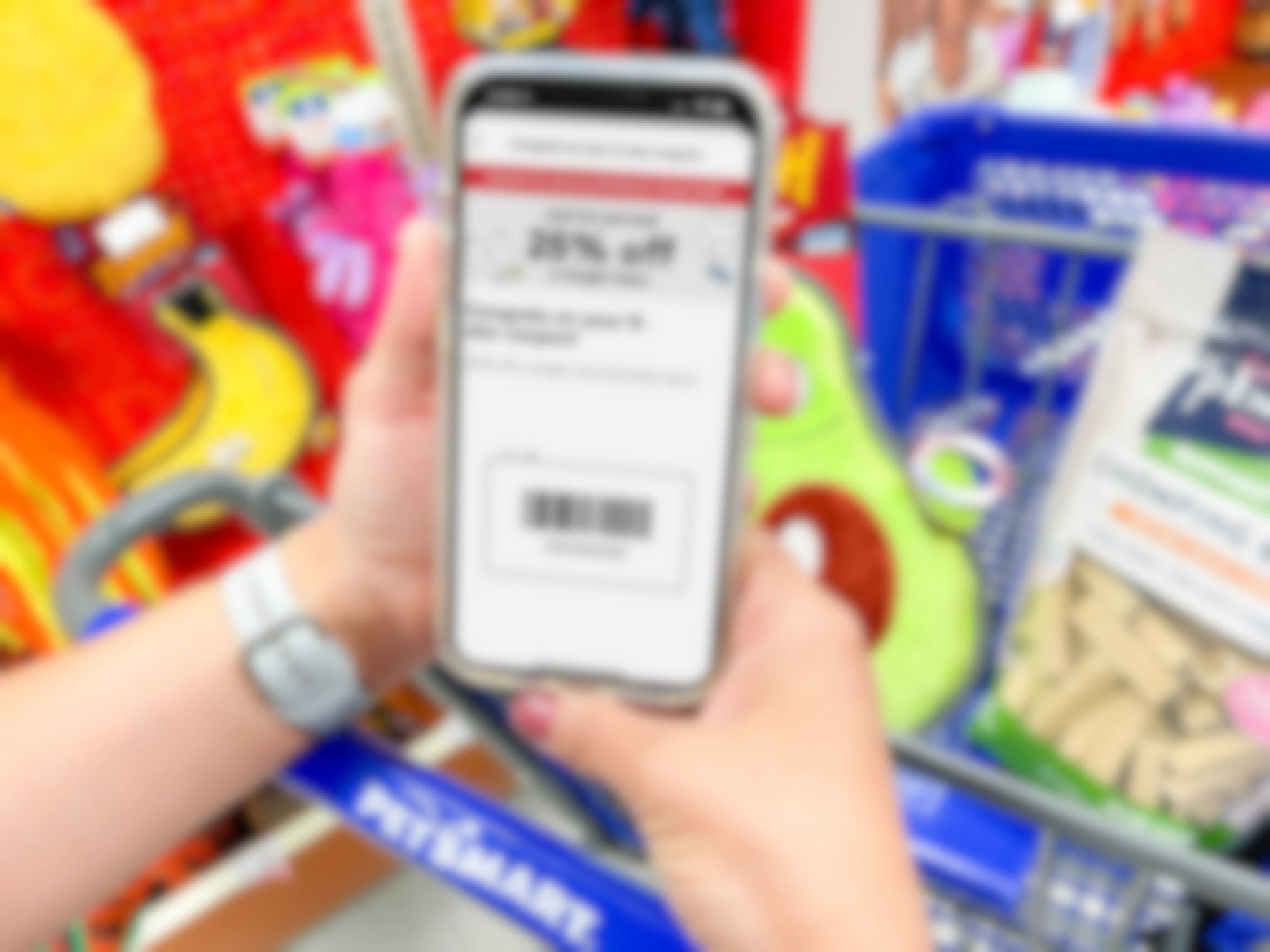 A person's hands holding a cellphone displaying a Treat Trail game coupon on the Petsmart app in front of a PetSmart shopping cart with a dog toy and treats in the basket.