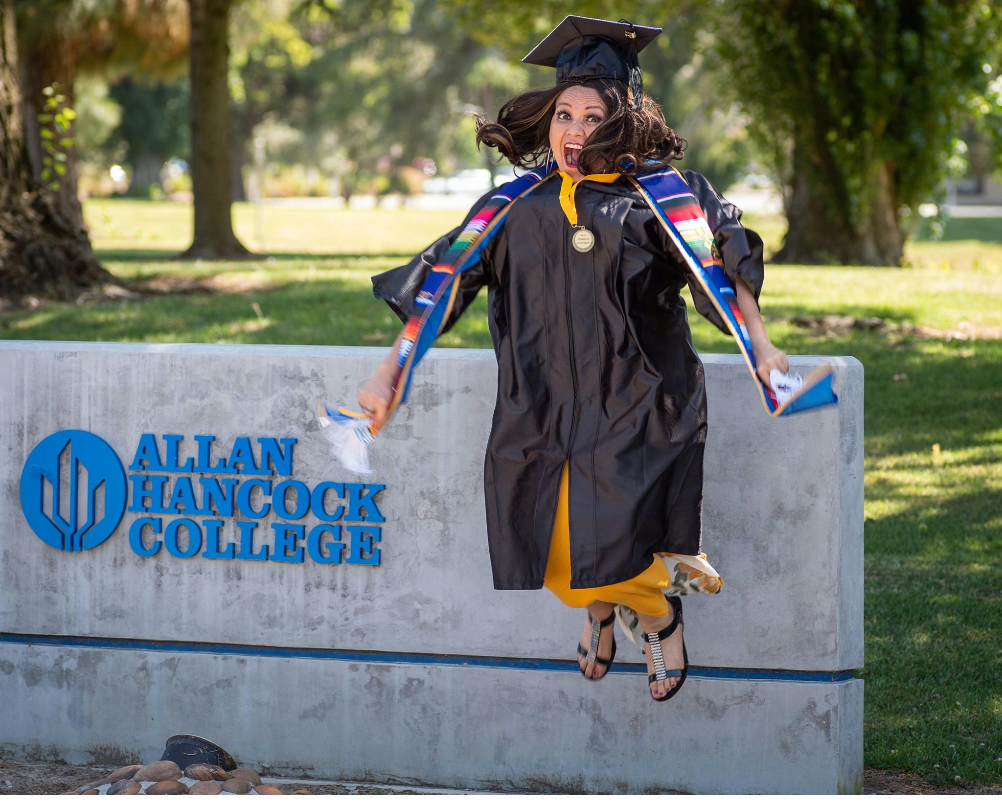 A student in a graduation cap and gown jumping up into the air next to a stone sign for Allan Hancock College.