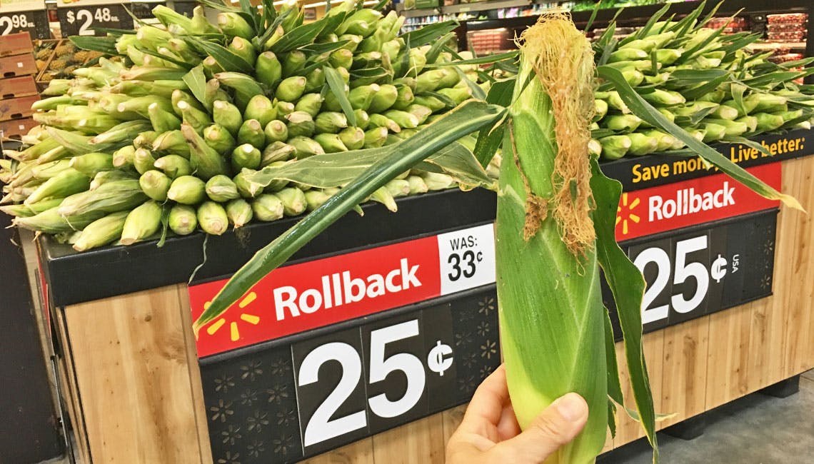 Hand holding an ear of corn in front of a Walmart Rollback sign that says 25 cents.