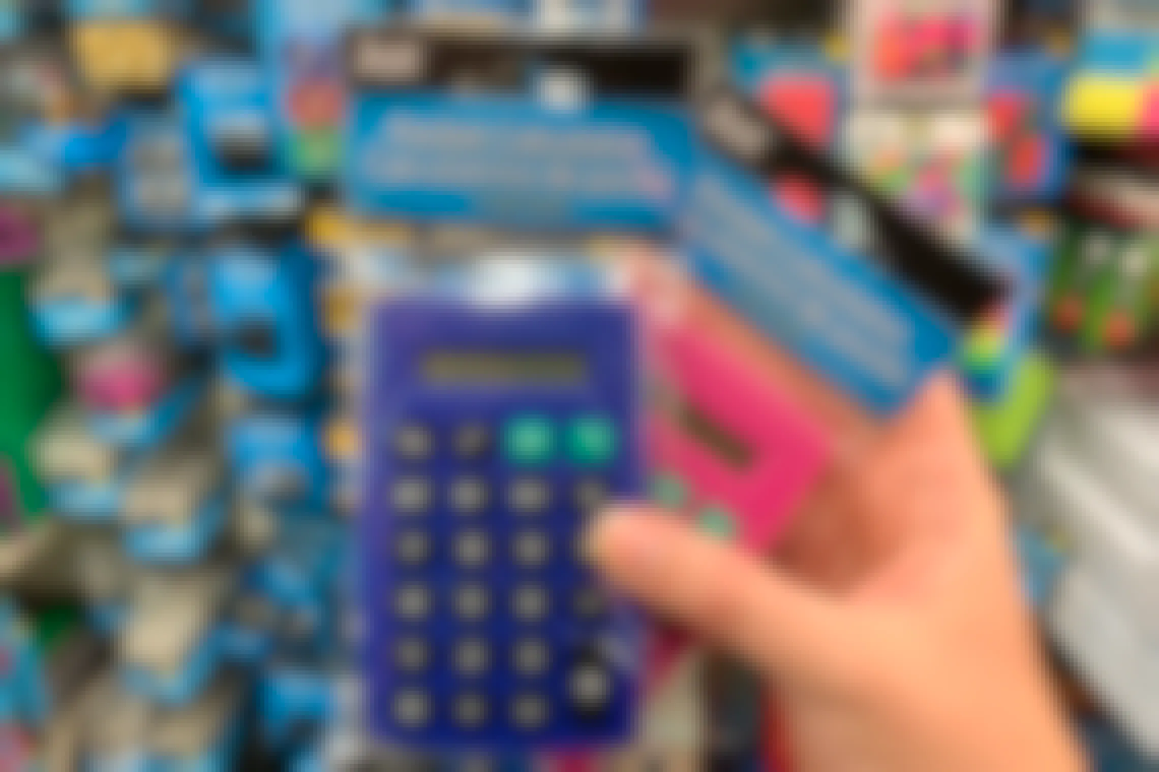A person's hand holding two Pocket Calculators in front of a shelf inside Dollar Tree.