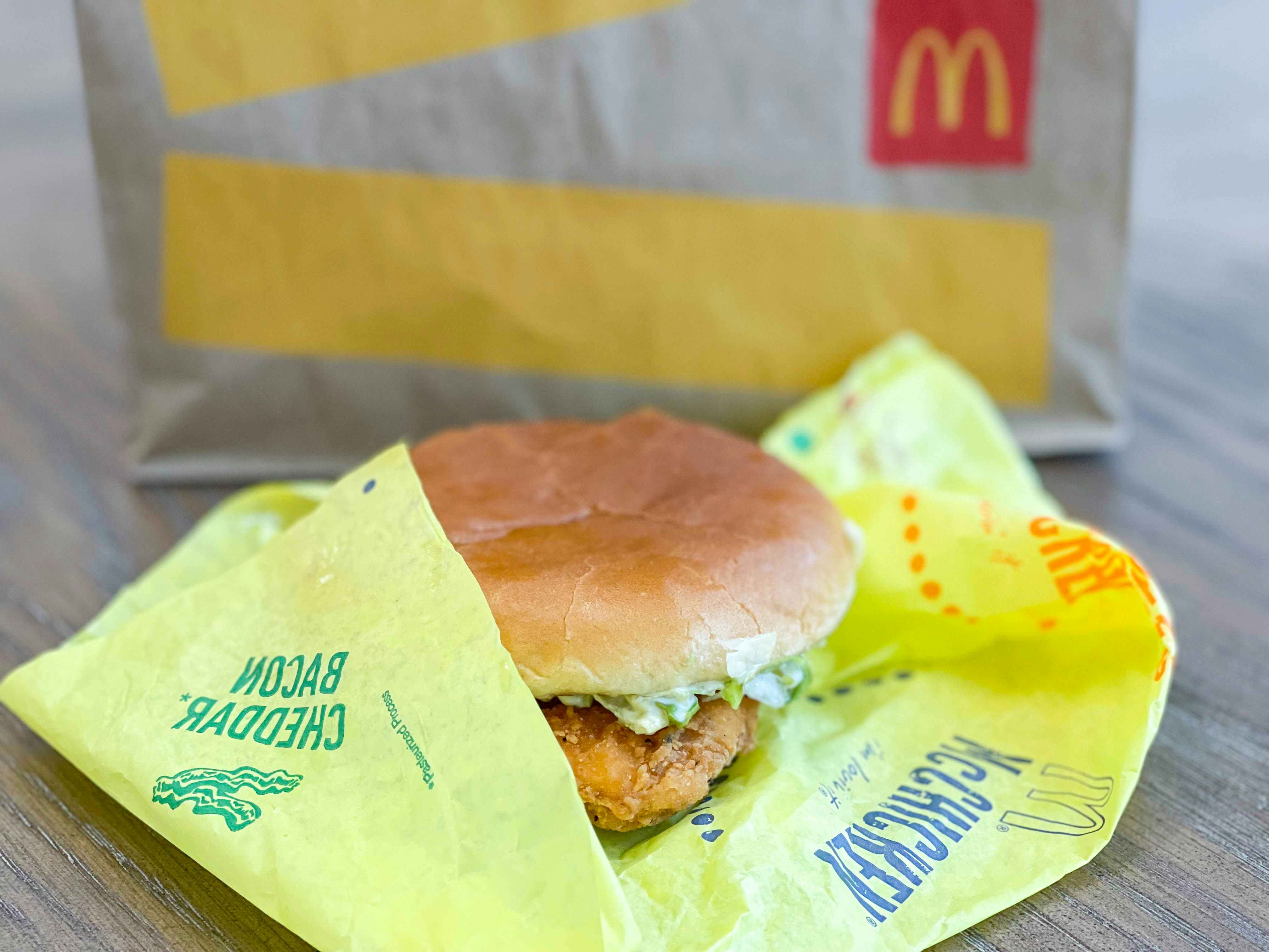 A close-up of a McChicken with bacon and cheese sitting on its paper wrapper in front of a McDonald's takeout bag.