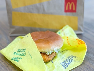 A close-up of a McChicken with bacon and cheese sitting on its paper wrapper in front of a McDonald's takeout bag.