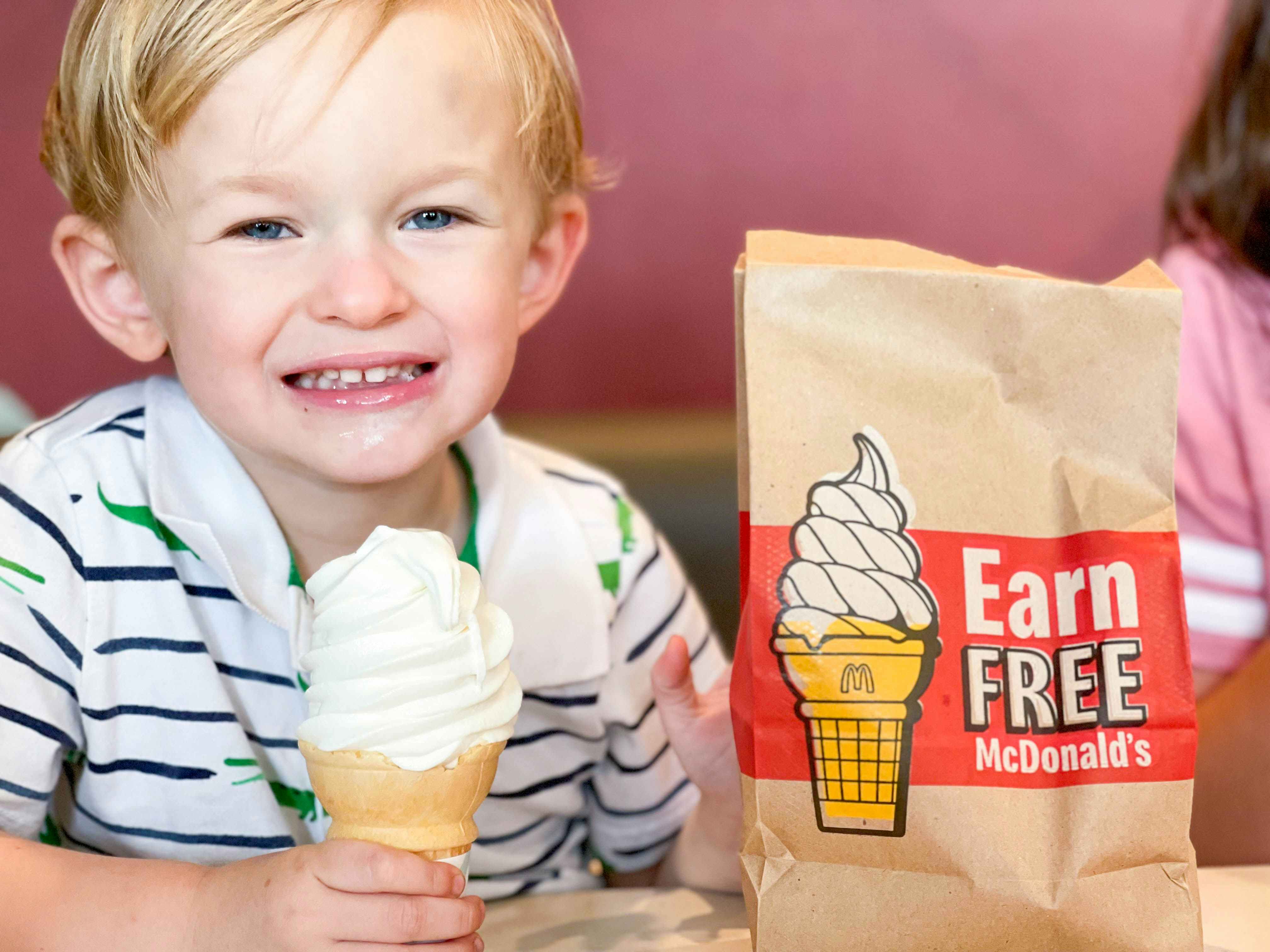 A child sitting in a booth at McDonald's, holding a soft serve ice cream cone next to a takeout bag that says, "Earn Free McDonald's