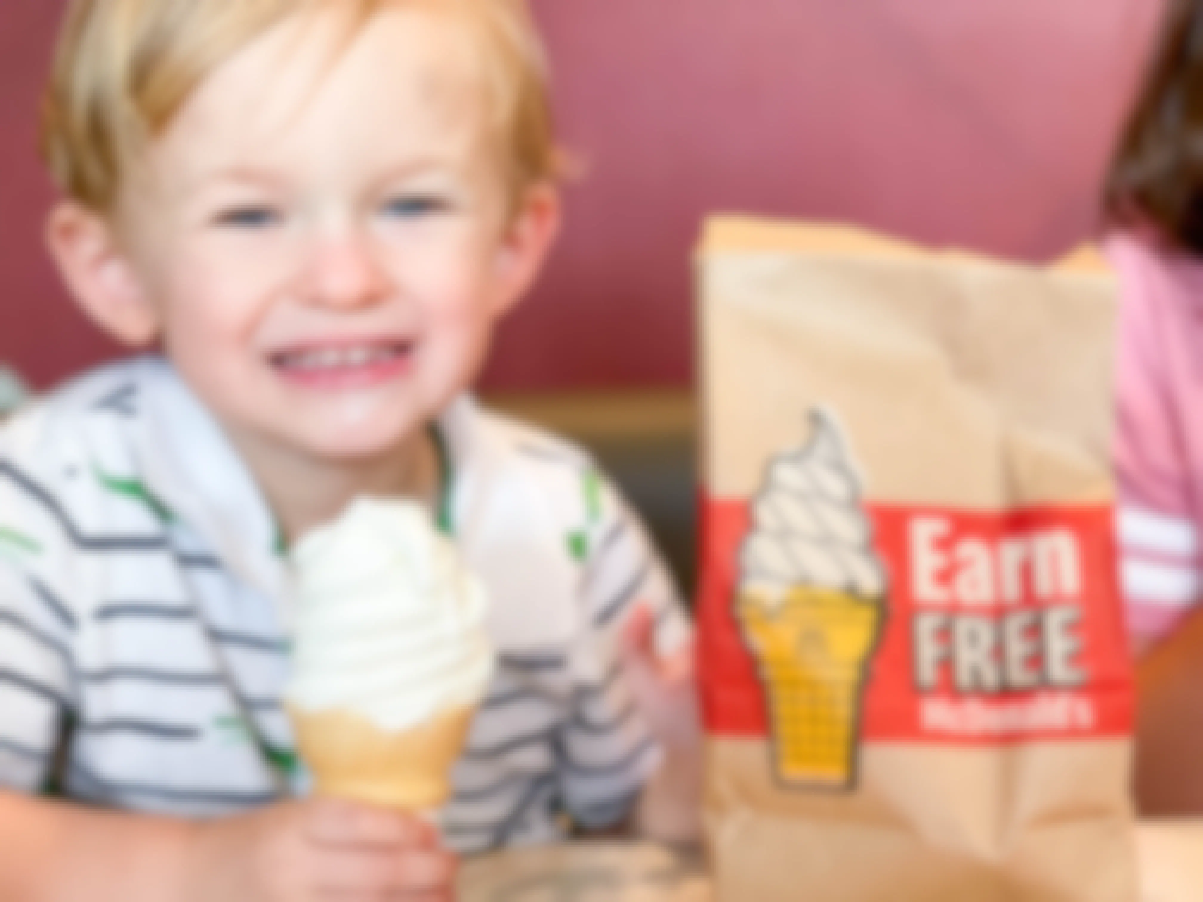 A child sitting in a booth at McDonald's, holding a soft serve ice cream cone next to a takeout bag that says, "Earn Free McDonald's