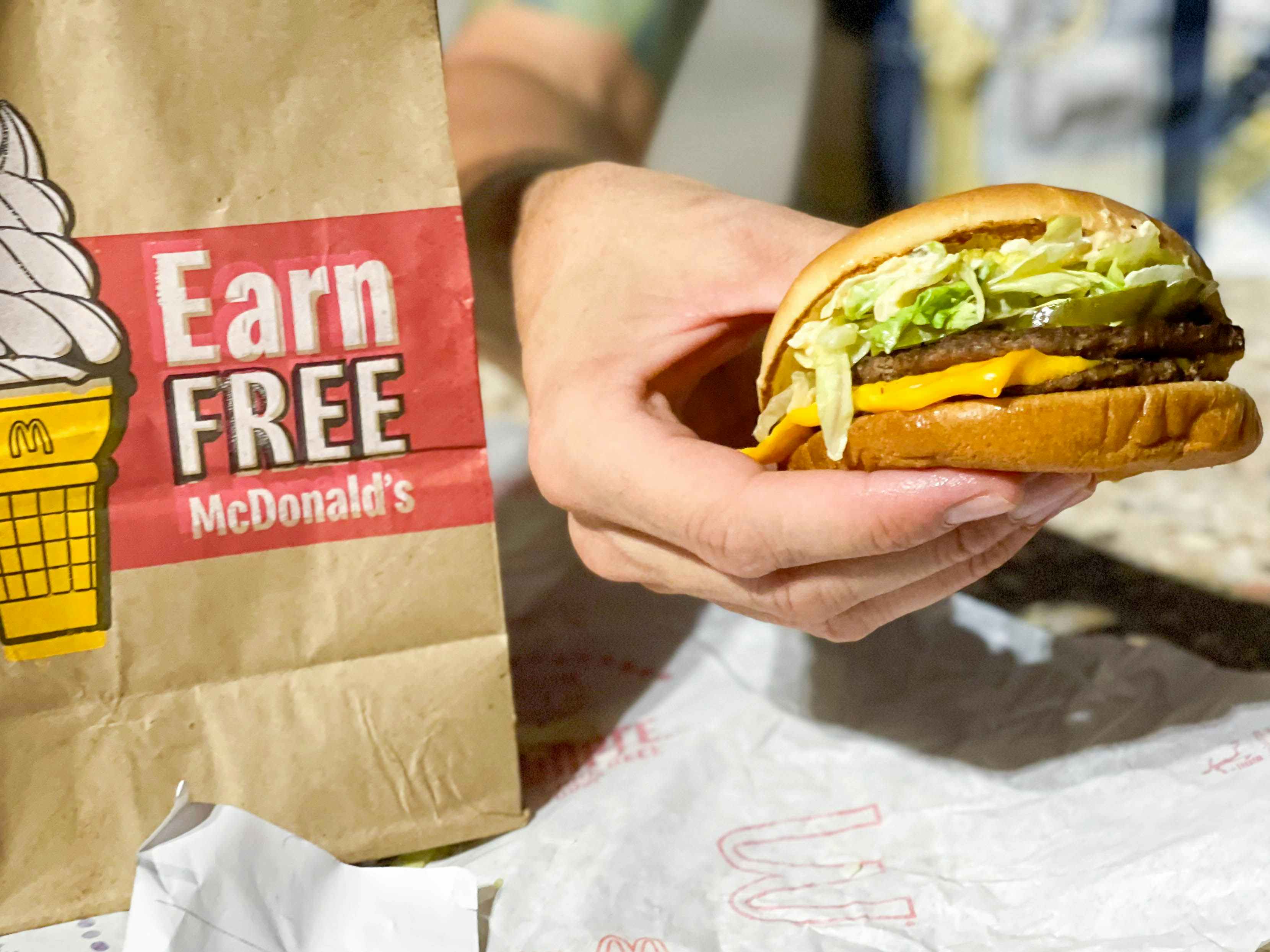 A person holding a McDouble special order burger from McDonald's next to a McDonald's takeout bag that says "Earn FREE McDonald's
