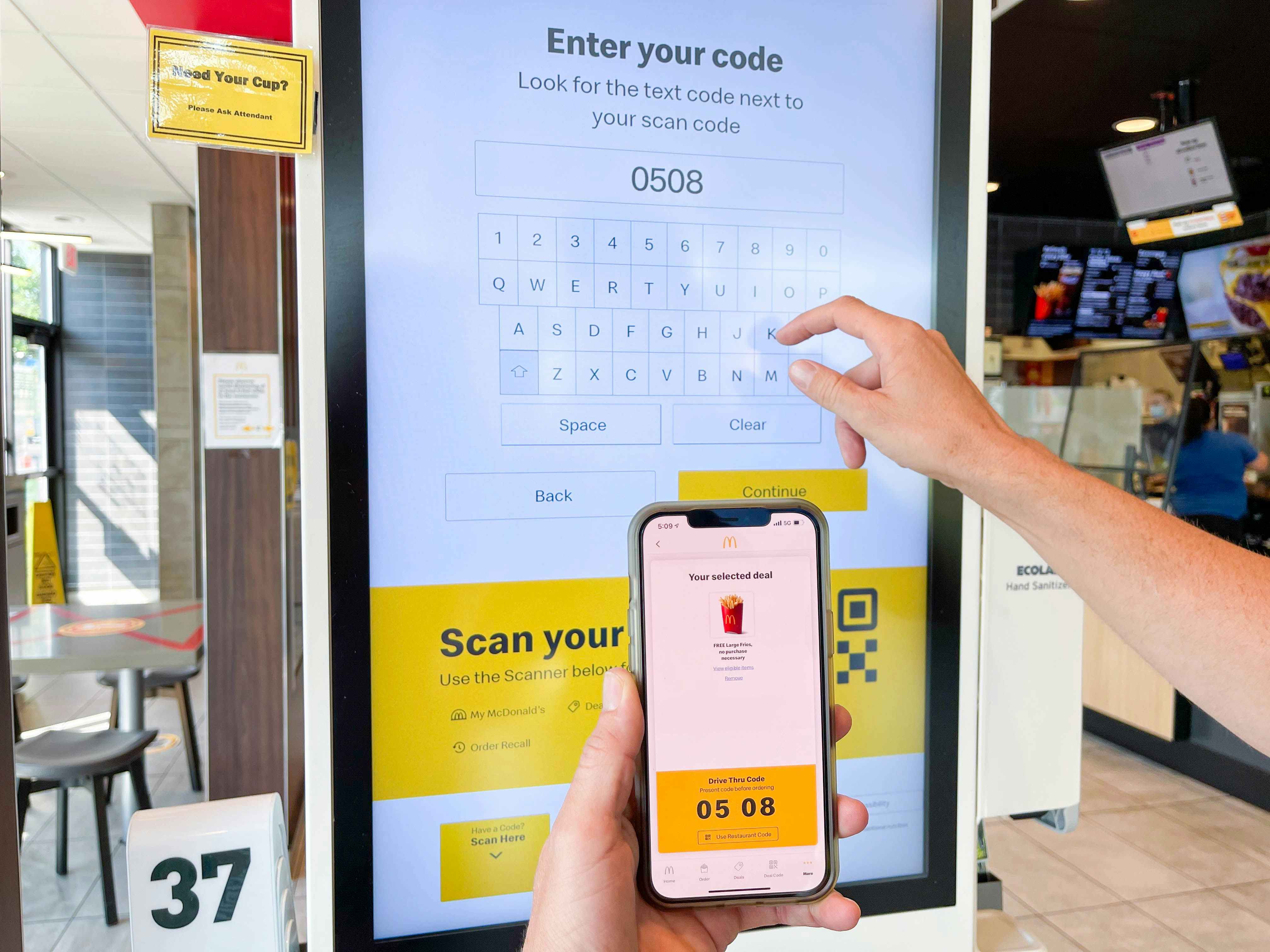 A person holding their phone displaying the McDonald's app code and typing it in on a big ordering screen inside McDonald's.