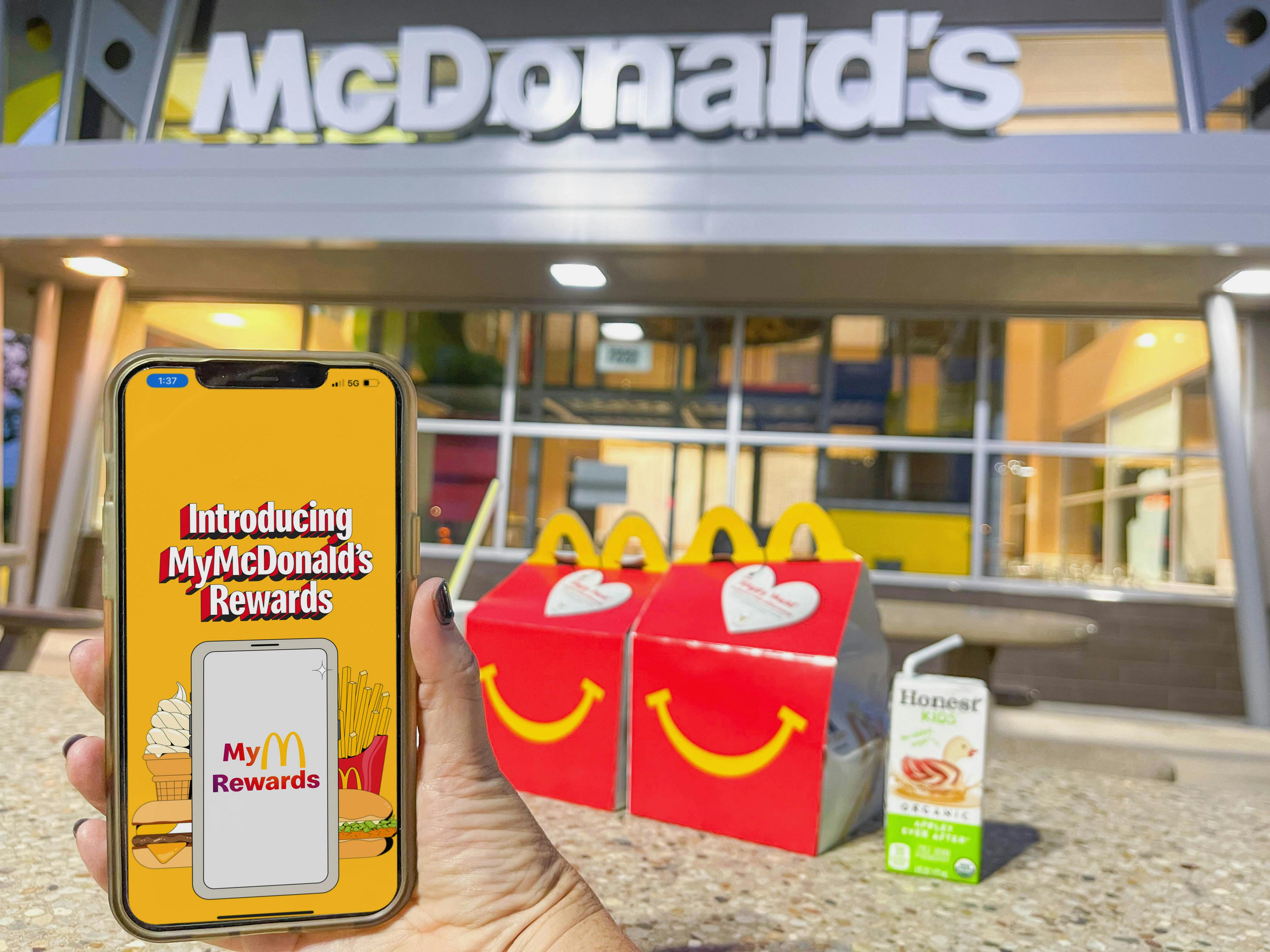 A person's hand holding up a cellphone displaying the McDonald's Rewards app in front of two Happy Meals and a juice box sitting on a table outside of a McDonald's restaurant.