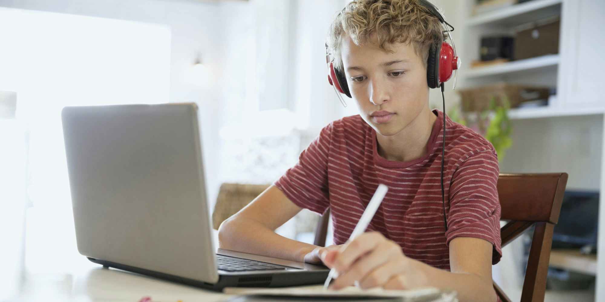 A teenager with headphones on sitting in front of a laptop and writing on a notepad.