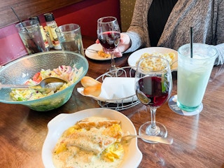 A person sitting at a table in Olive Garden with plates of food, drinks, breadtsticks, and a salad bowl.