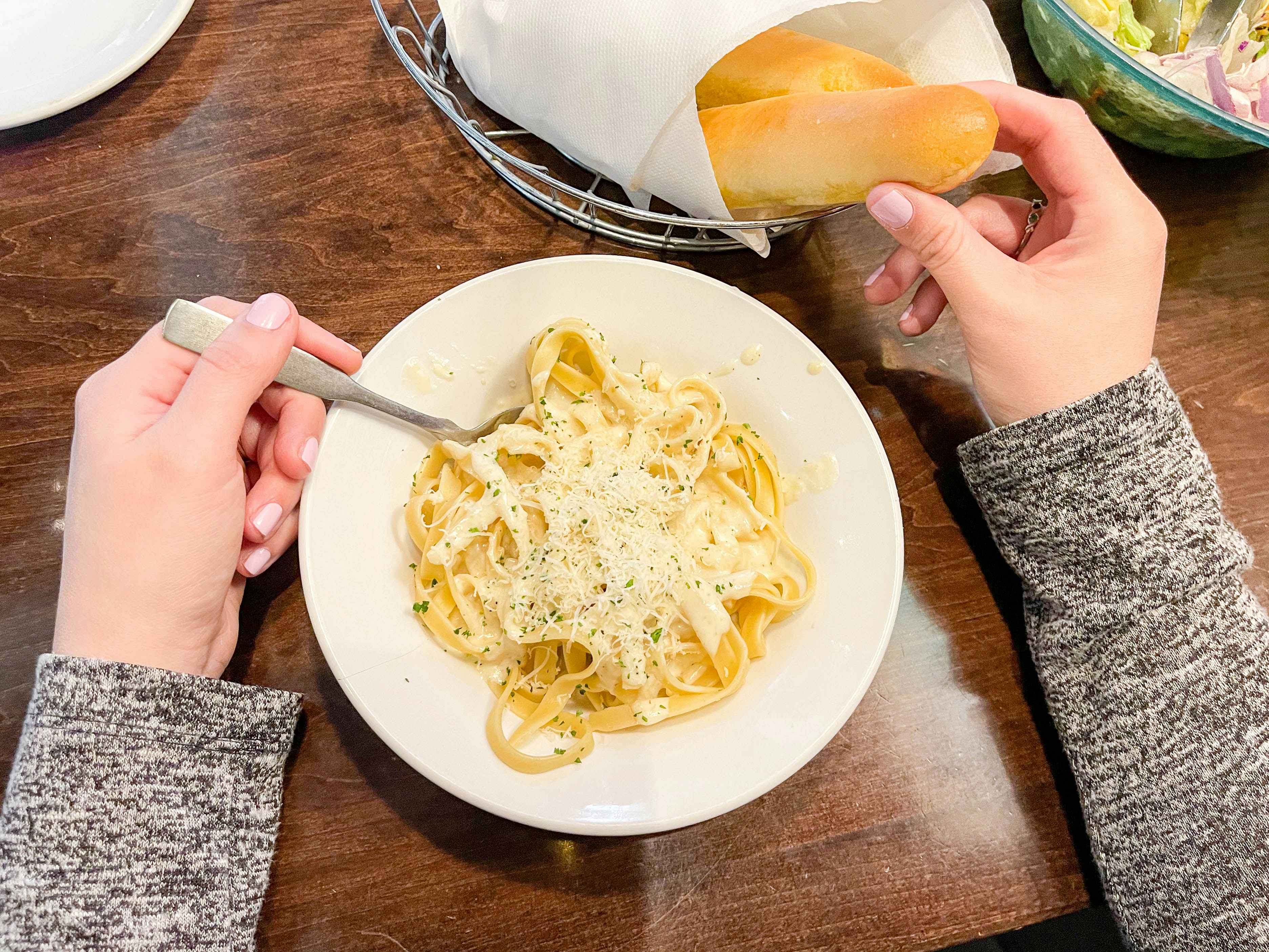 A person sitting at a table at Olive Garden, taking a breadstick from the bread basket and putting a fork into a bowl of pasta.