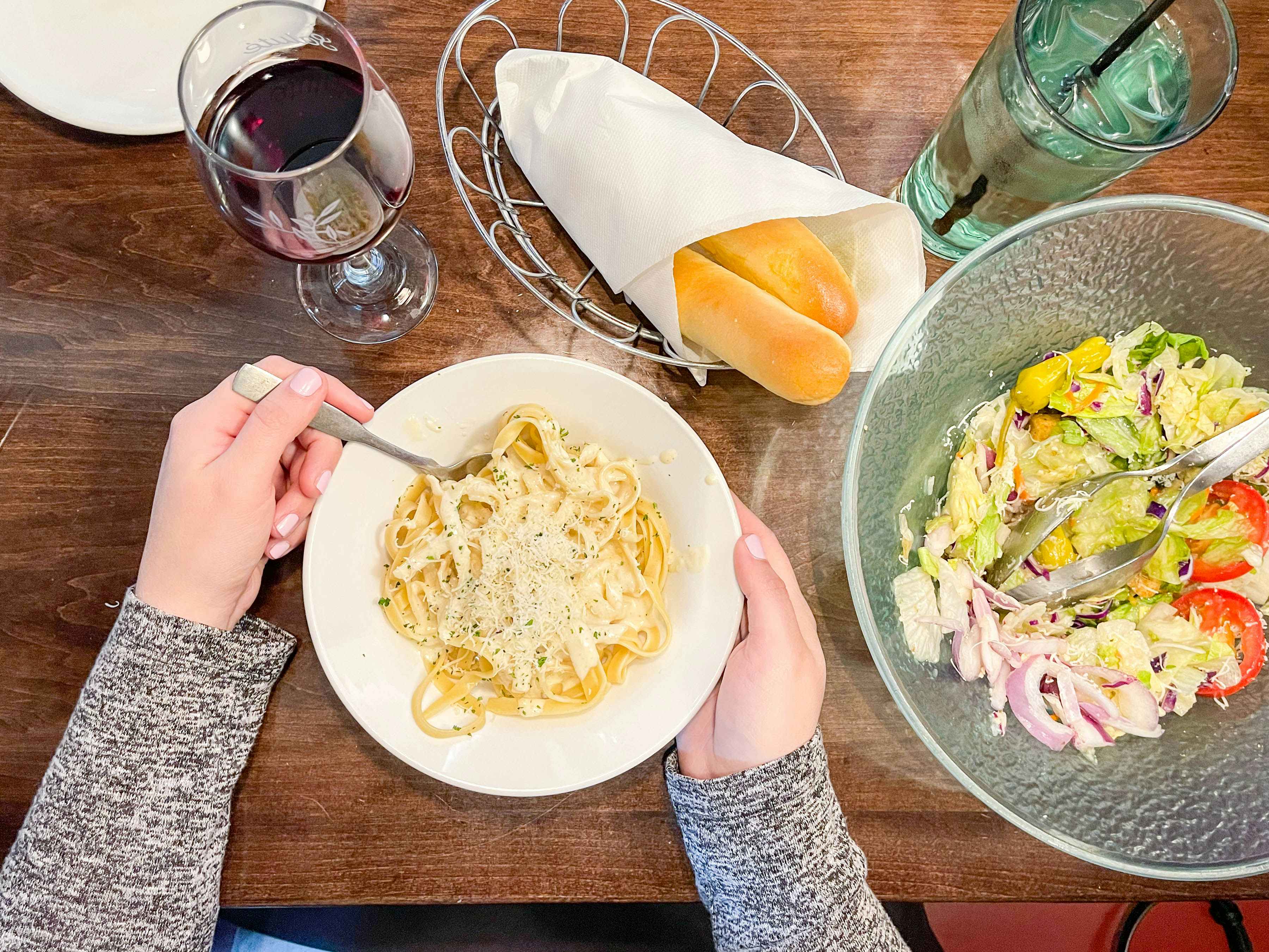 A person sitting at Olive Garden with a bowl of pasta, a bowl of salad, a basket of breadsticks, and a glass of wine on the table in front of them.