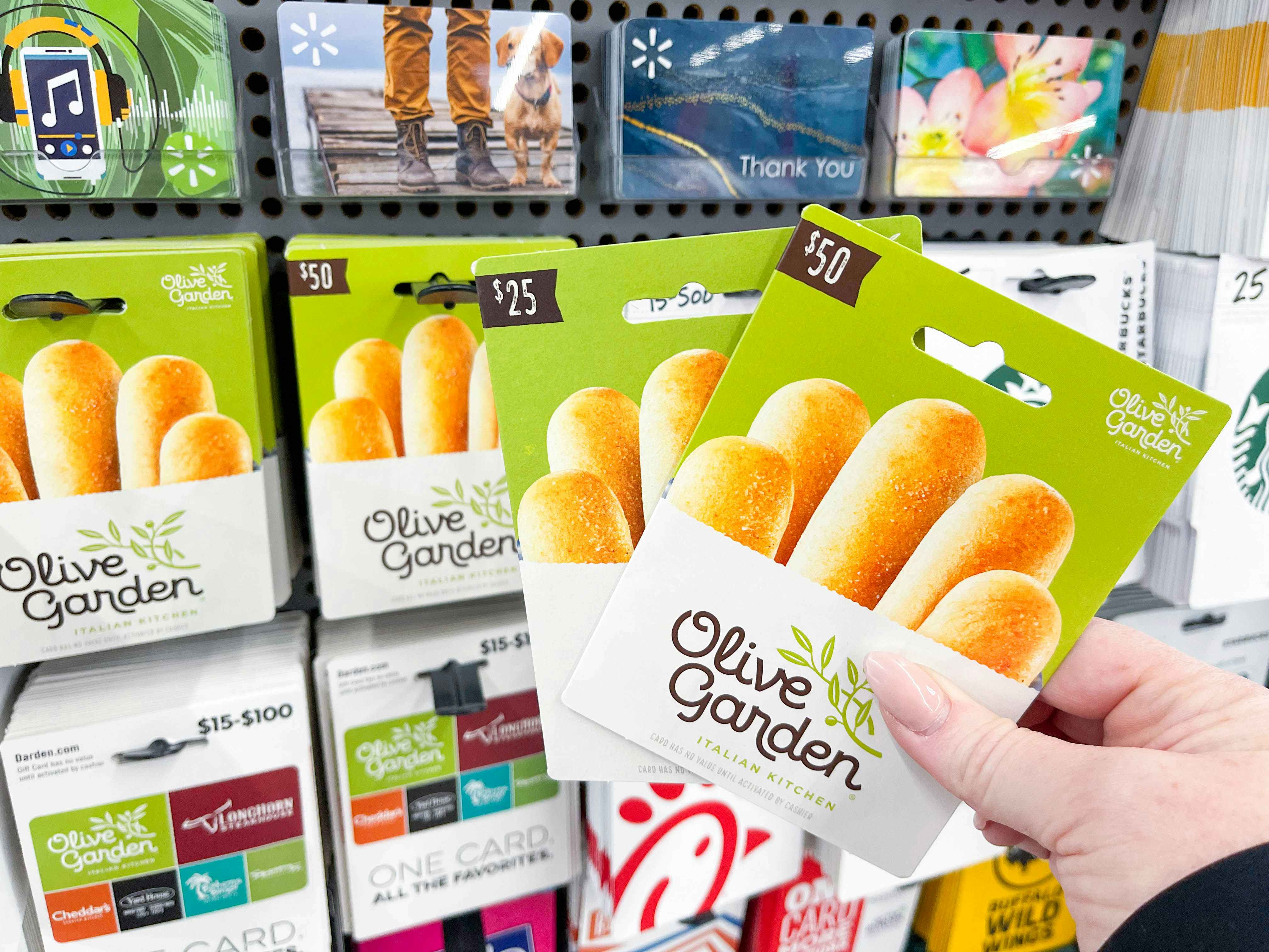 a person holding two olive garden gift cards in front of a lot of gift cards in store display 