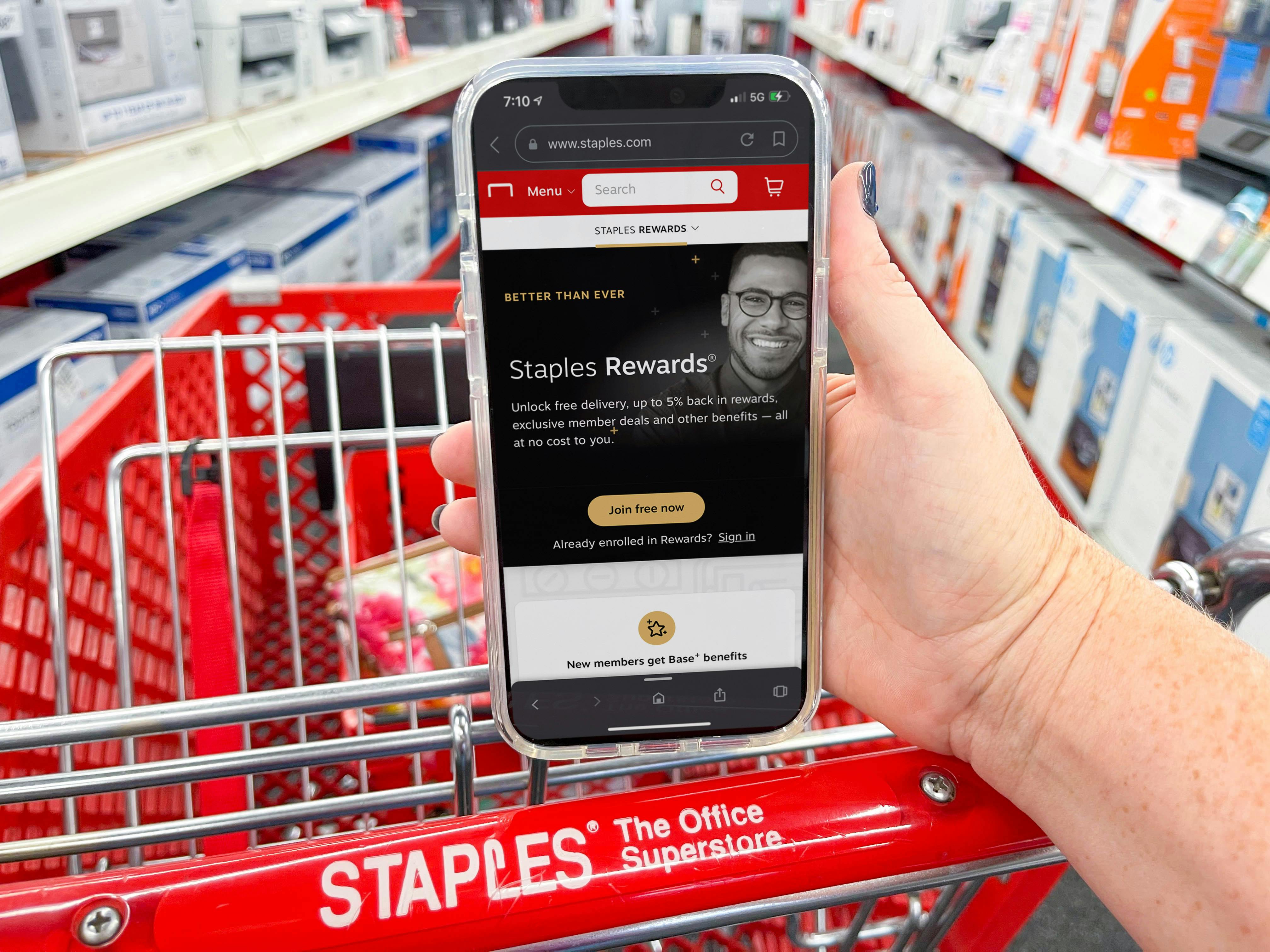 A person's hand resting on the handle of a Staples shopping cart, holding a cell phone displaying the Staples Rewards page on their website.